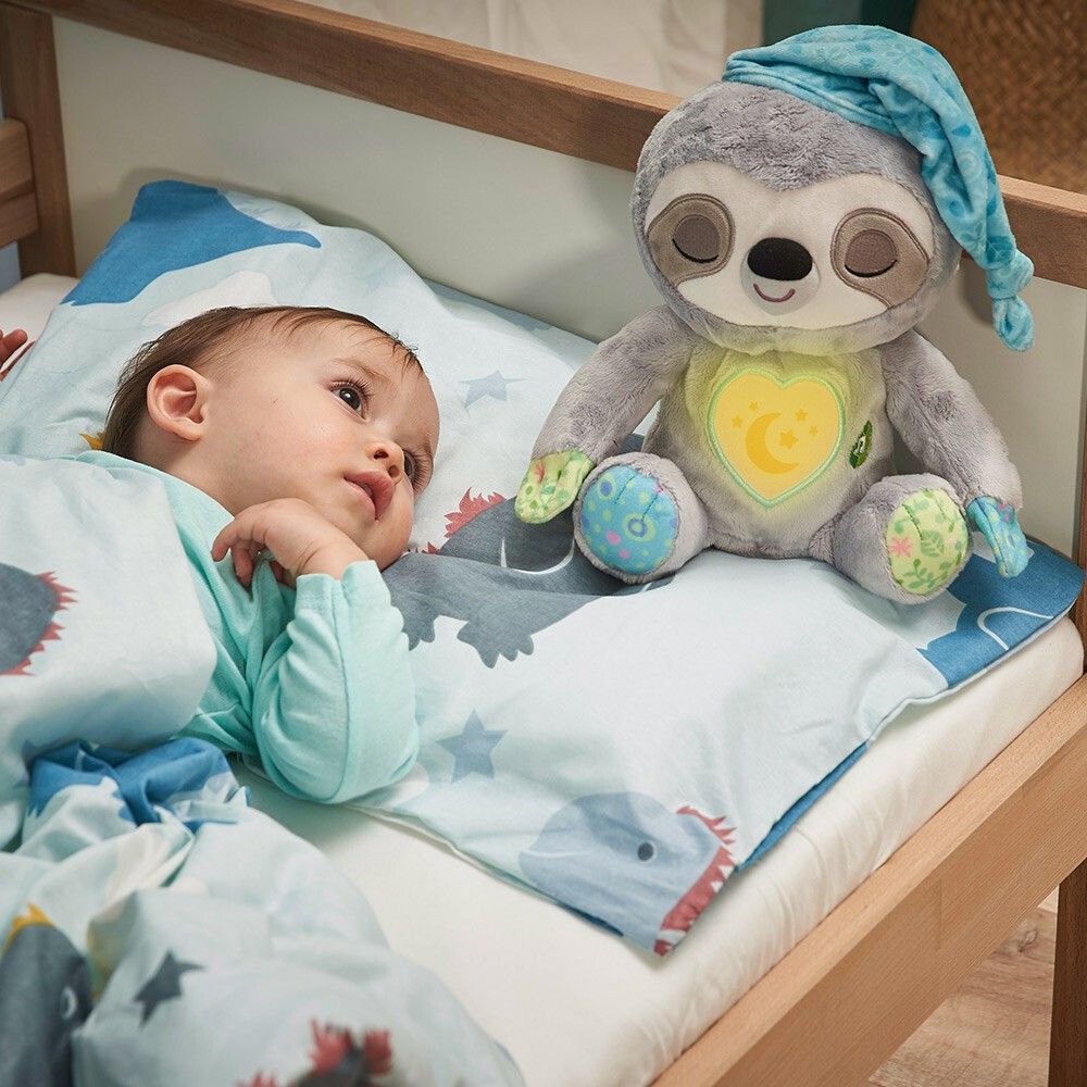 Bambino a letto accanto a un peluche bradipo grigio con cappello. Cuore giallo con motivo luna-stella.