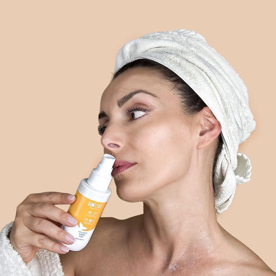 Donna con asciugamano tiene flacone spray bianco. SUNCHIÒ AIR-FIT SPF50+ visibile. Etichetta gialla.