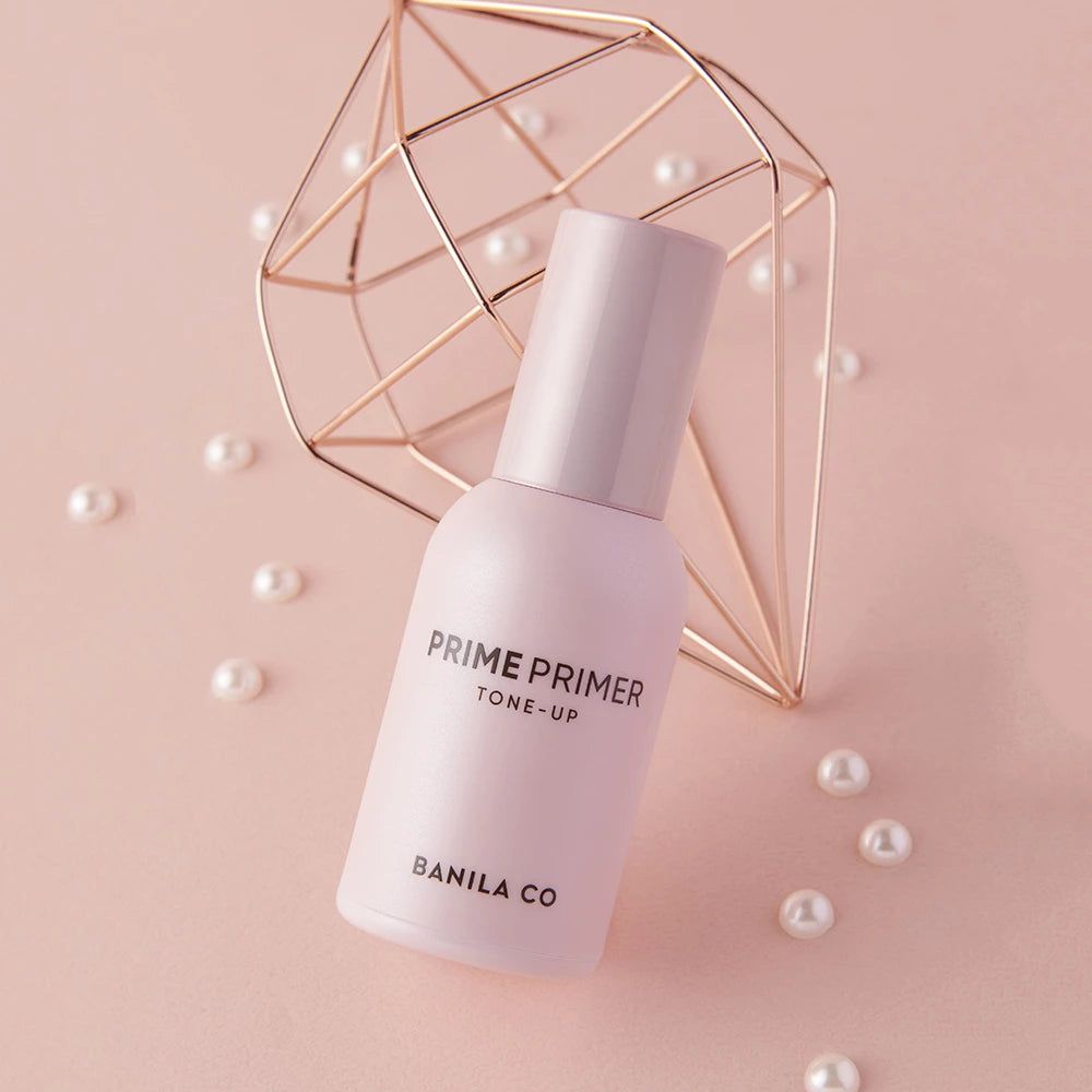 Flacone di primer rosa con tappo. Scritta: PRIME PRIMER TONE-UP, BANILA CO. Decorazione con perle e forma dorata.