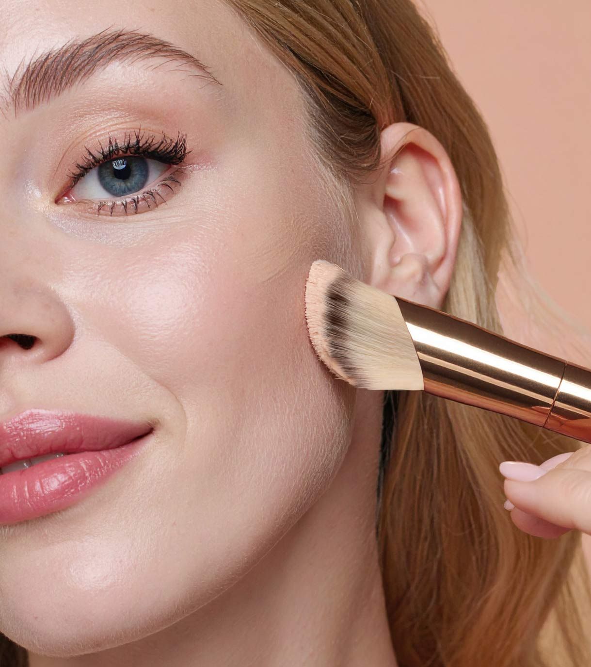 Un pennello da trucco con manico dorato e testa angolata viene applicato sulla guancia di una donna.