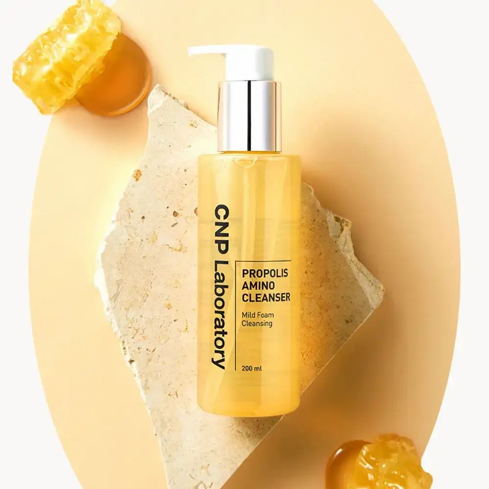 Flacone giallo con pompa. Scritta: CNP Laboratory, Propolis Amino Cleanser. Posizionato su pietra. Favo e gocce.