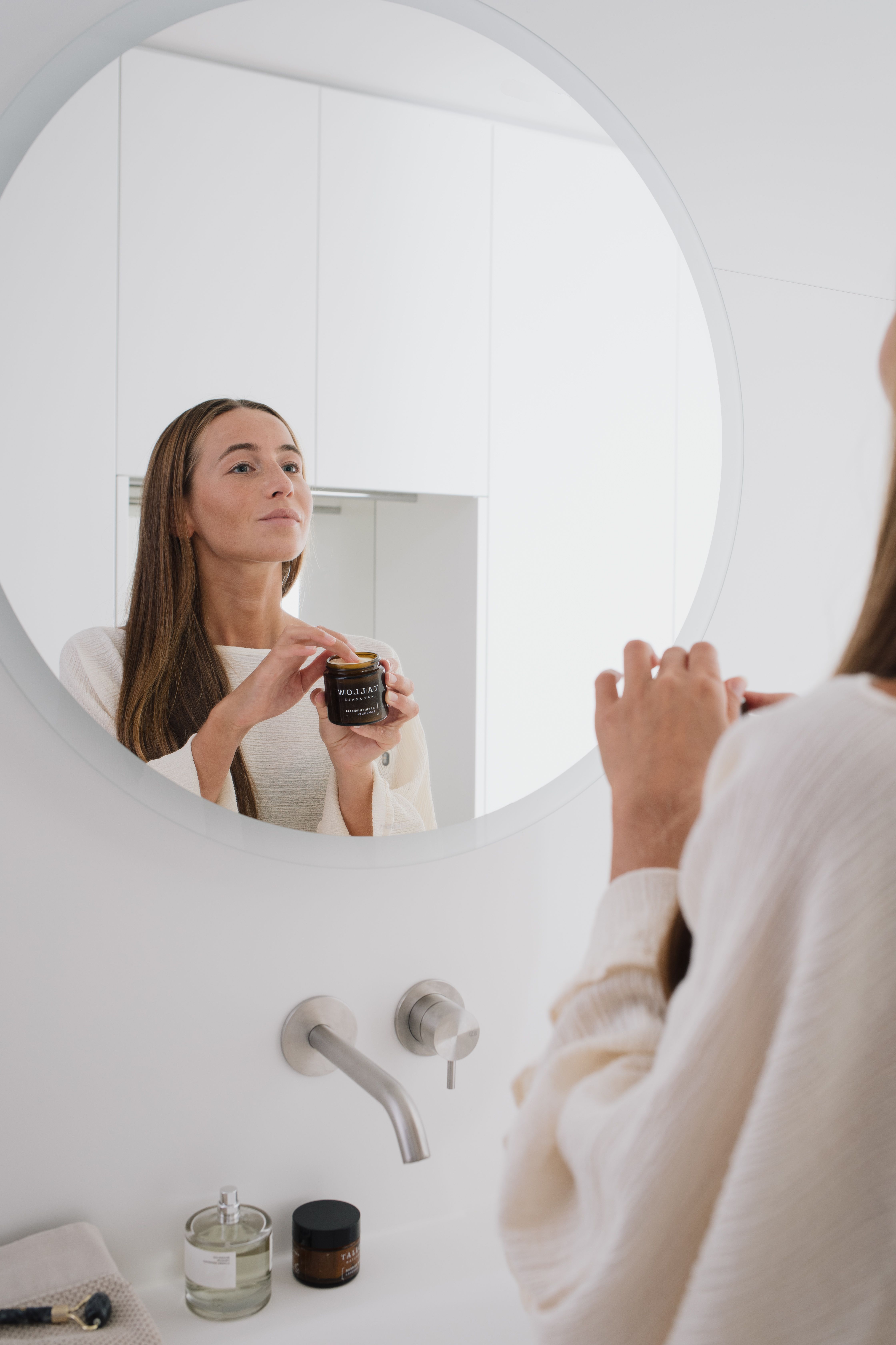 Donna si guarda allo specchio. Tiene un barattolo marrone con coperchio nero. Scritta: Tallow Naturals, Barrier Repair Pur.