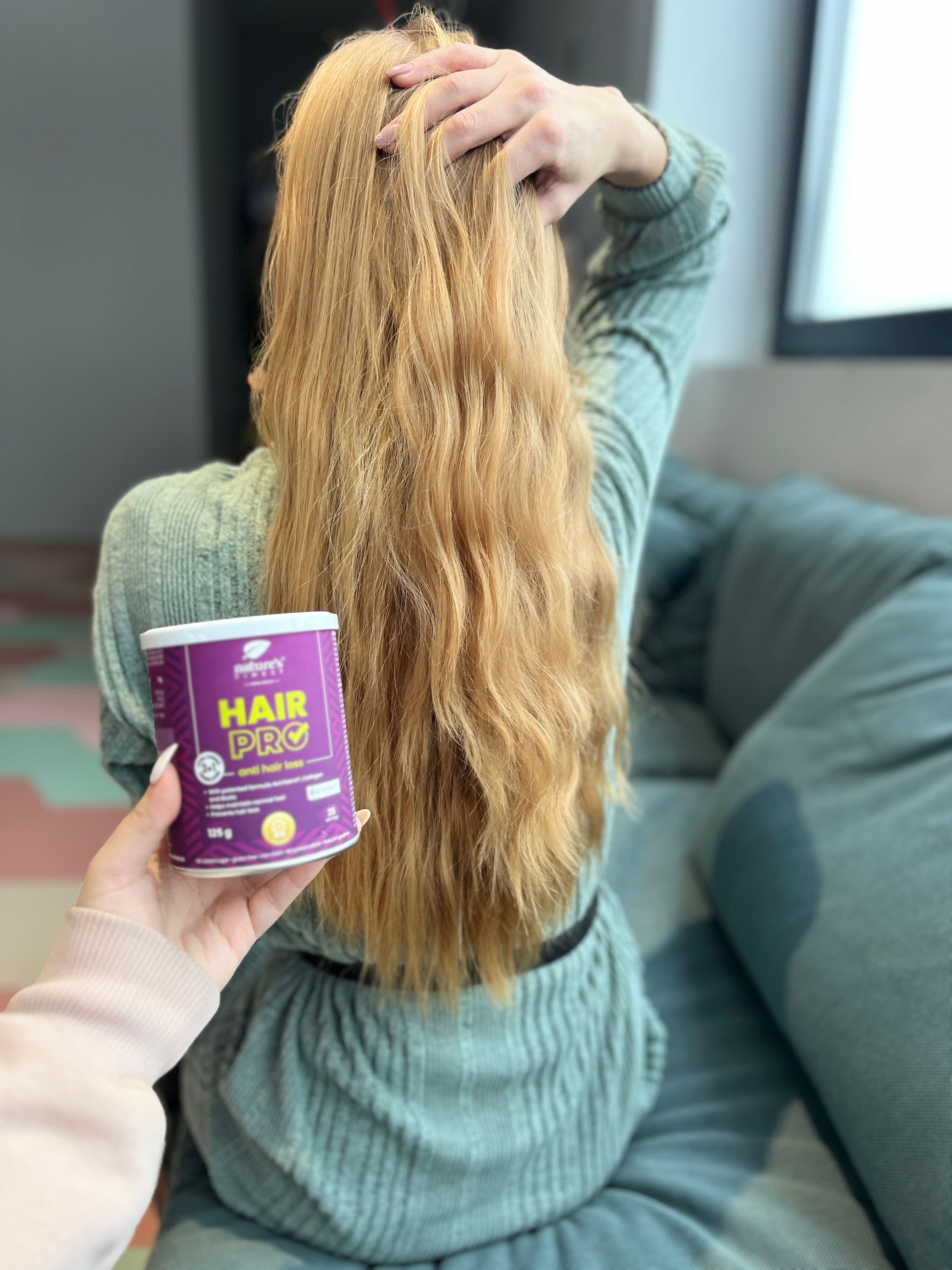 Persona tiene barattolo Nature's Finest Hair Pro. Scritta: Hair Pro, contro la caduta dei capelli. 125g, 25 porzioni. Capelli lunghi.