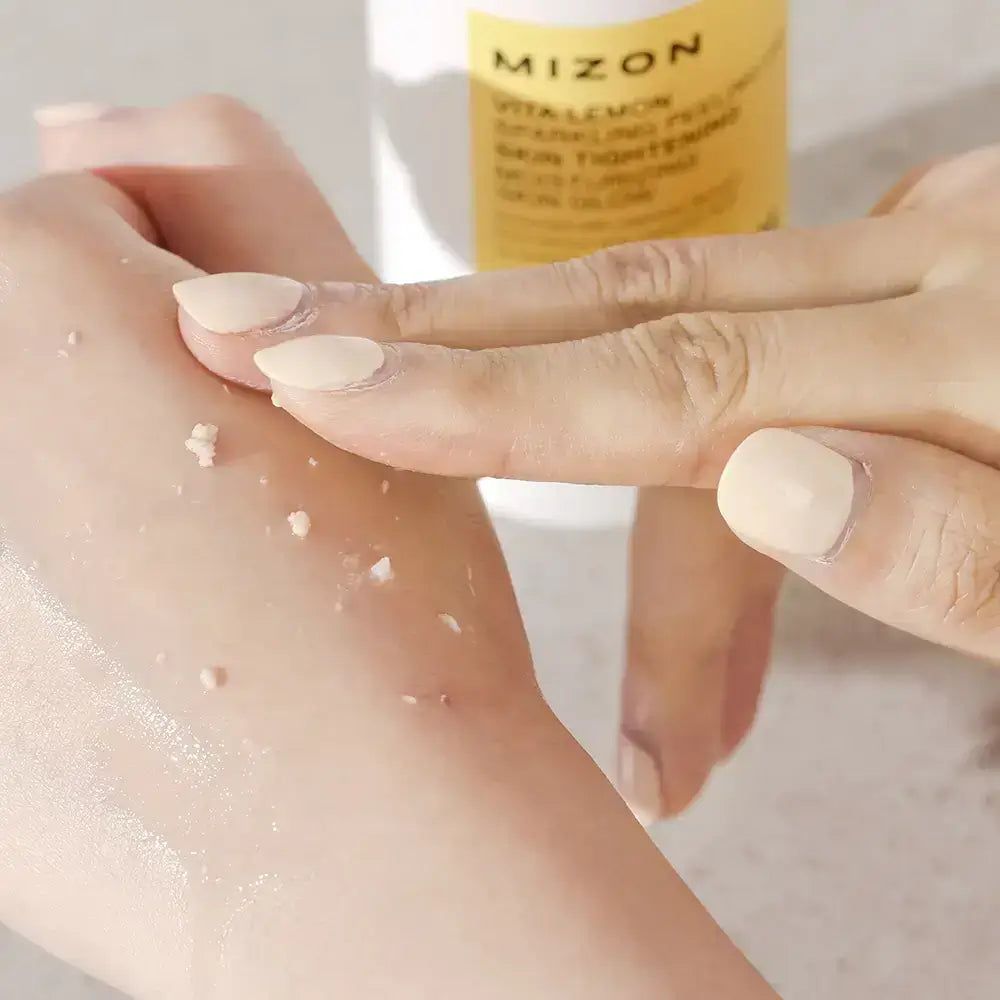 Mani che strofinano il gel sulla pelle. Residui di gel visibili. Prodotto sullo sfondo. MIZON Vita Lemon Sparkling Peeling Gel.