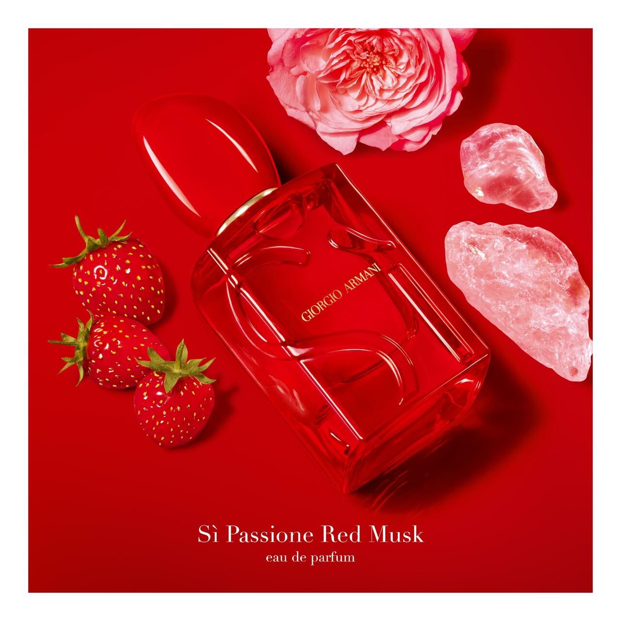Flacone rosso Giorgio Armani Sì Passione Red Musk, circondato da fragole, rosa e pietre su sfondo rosso.