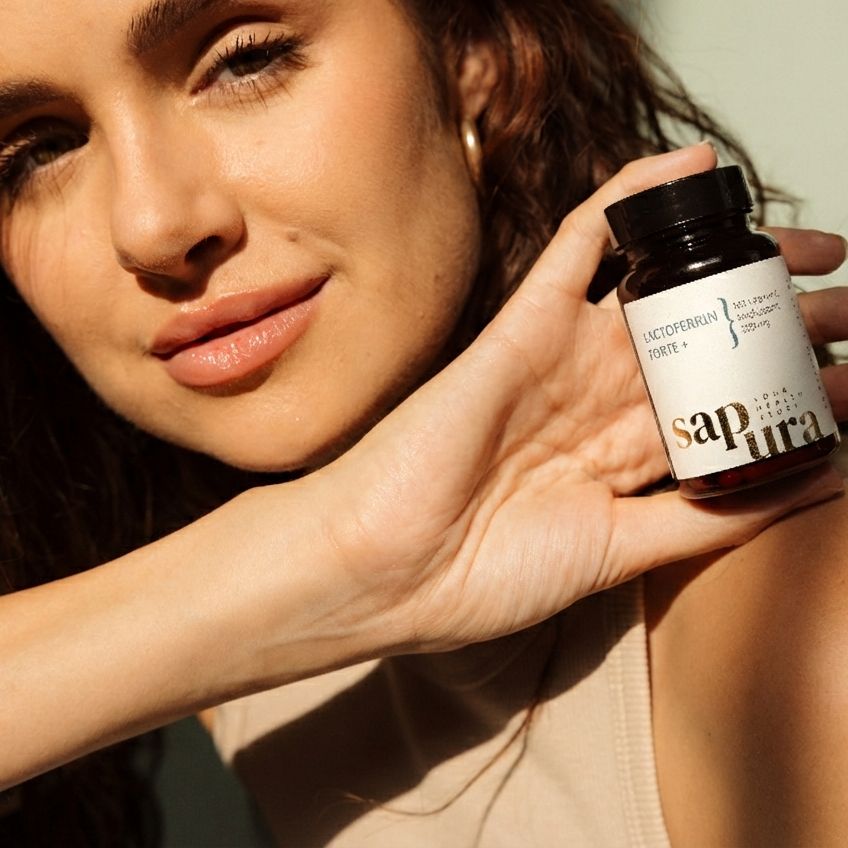 Donna tiene in mano un flacone marrone. Scritta: Lactoferrin Forte +, Vitamina C, Sapura. Flacone con tappo nero.