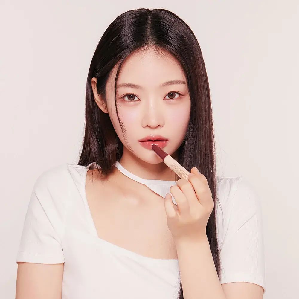 Giovane donna con lunghi capelli scuri e maglione bianco applica un lucidalabbra. Il rossetto è color prugna.