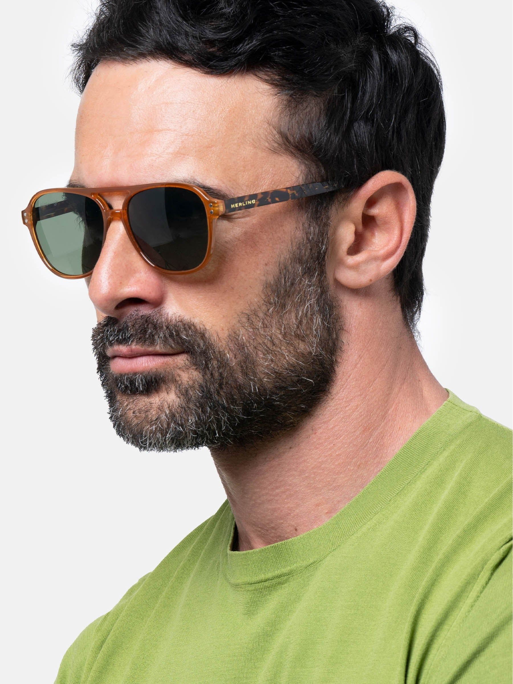 Uomo con occhiali da sole marroni e lenti verdi. Aste con motivo tartarugato. Barba.