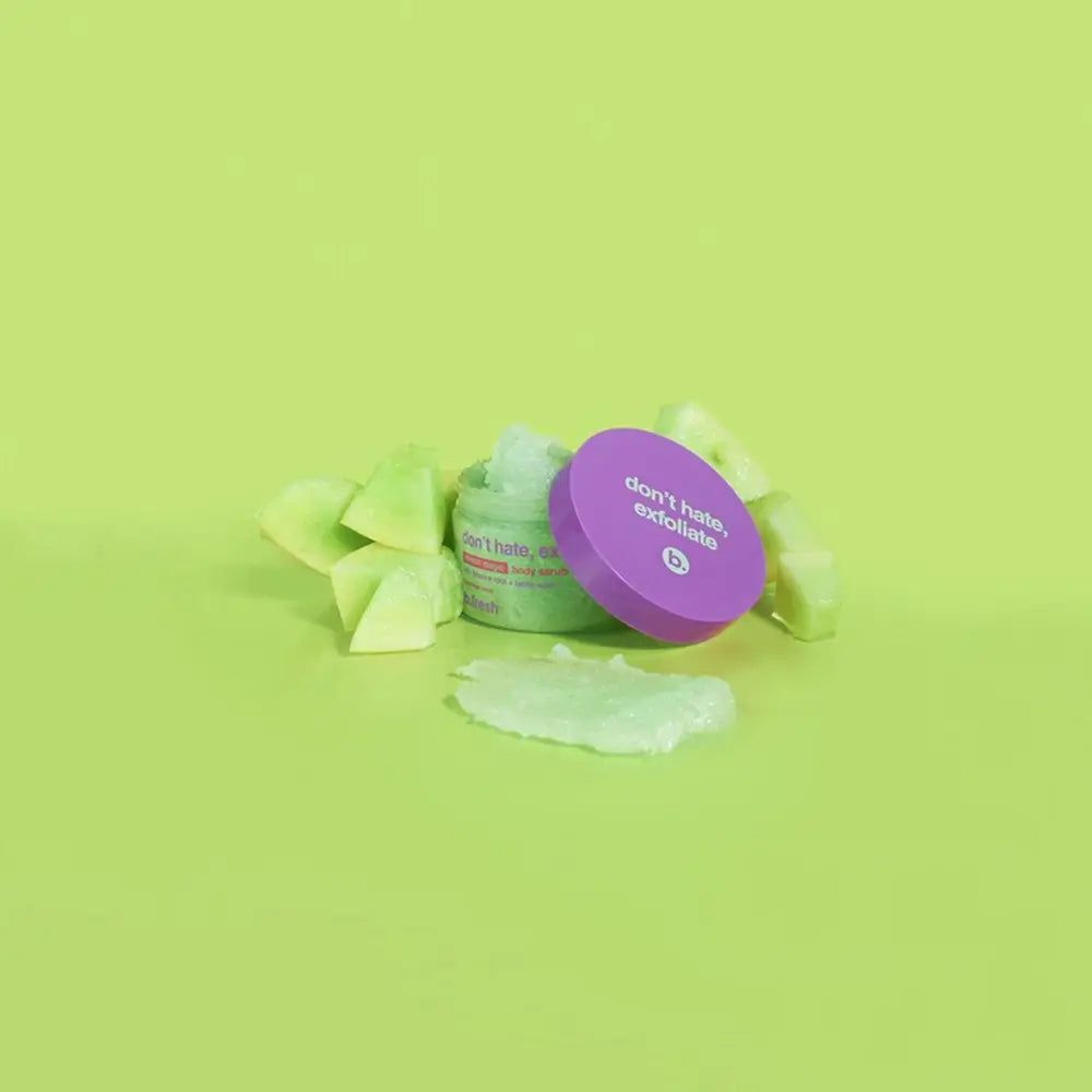 Contenitore verde per scrub esfoliante con coperchio viola, accanto a pezzi di frutta verde e scrub.