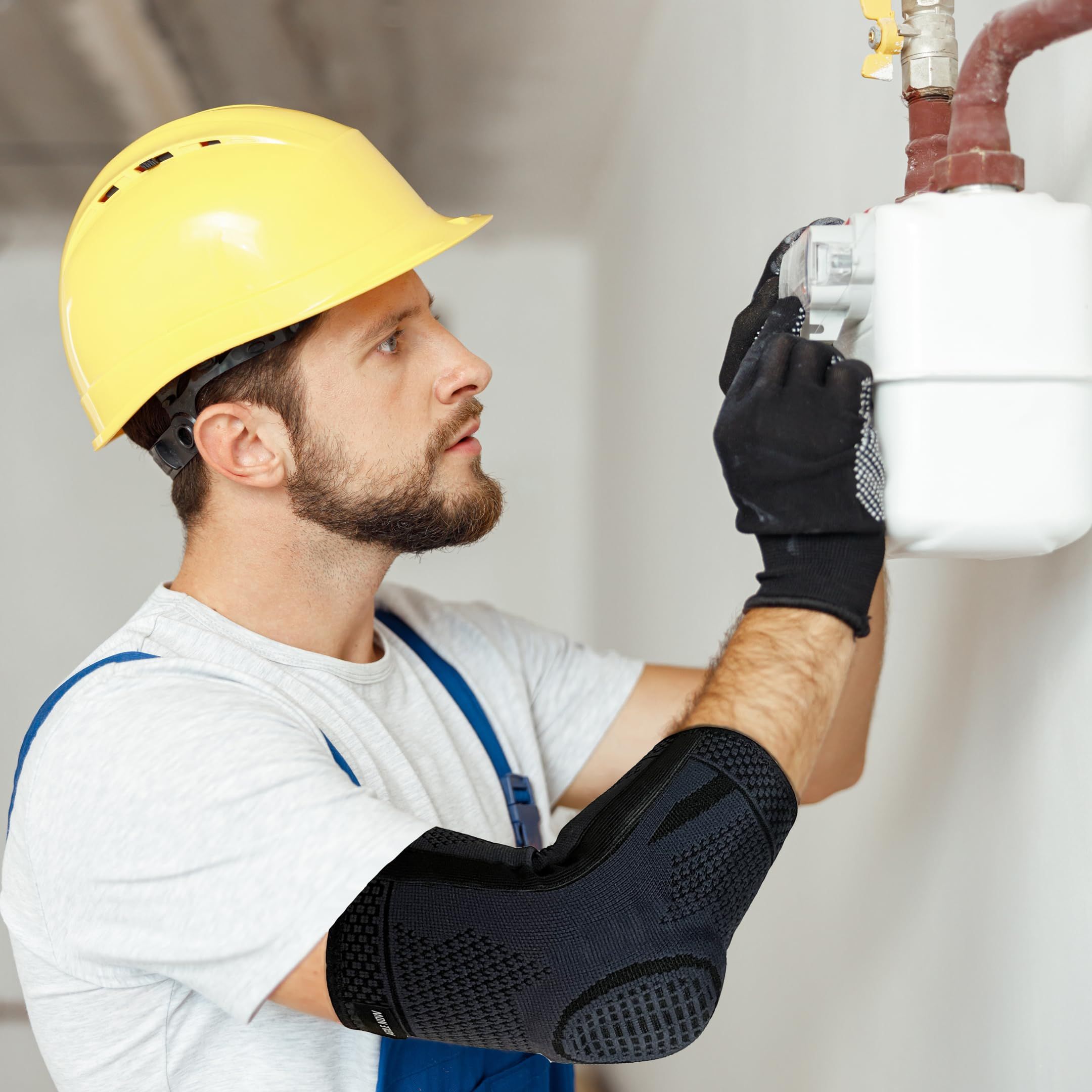 Artigiano con casco giallo e tutore per gomito nero. Sta lavorando su un contatore del gas.