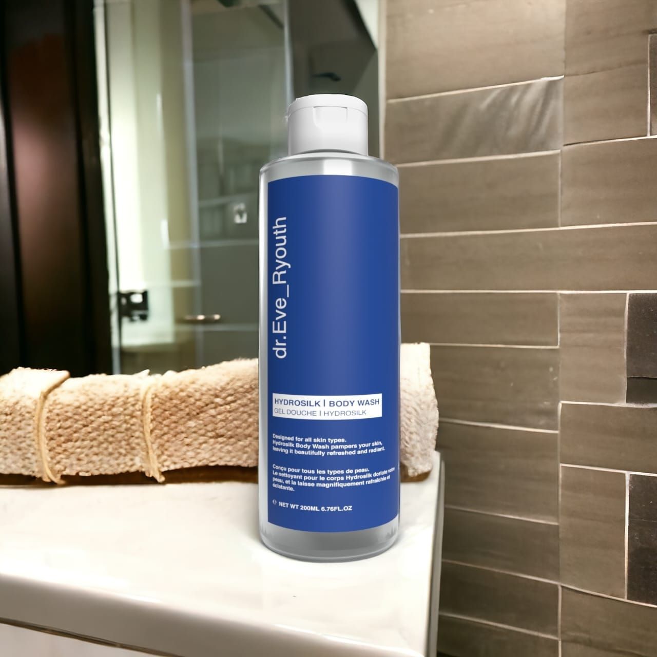 Flacone trasparente con etichetta blu. Scritta: dr.Eve_Ryouth, Hydrosilk Body Wash. Su una superficie bianca, asciugamano sullo sfondo.