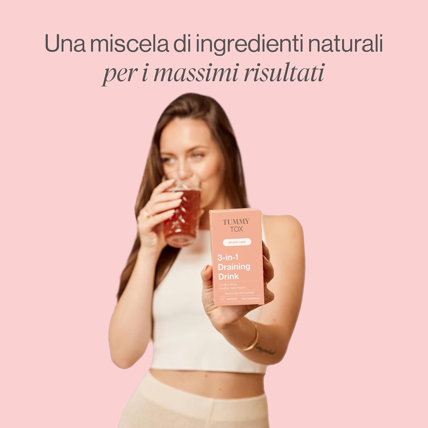 Donna che beve da un bicchiere. Tiene in mano una confezione con la scritta Tummy Tox 3-in-1 Draining Drink.