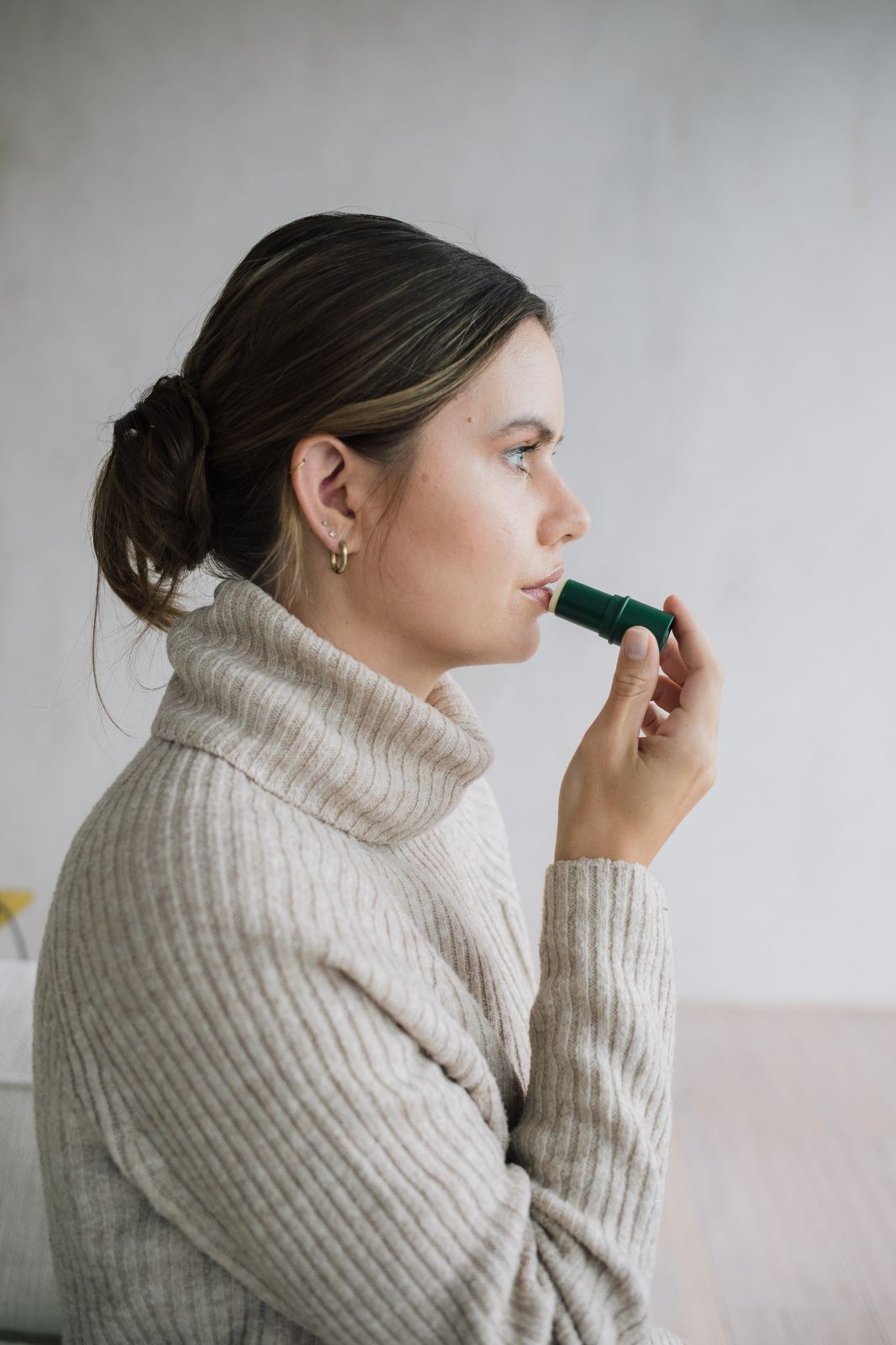 Donna che applica balsamo per labbra verde. Indossa un dolcevita beige. Vista di profilo.