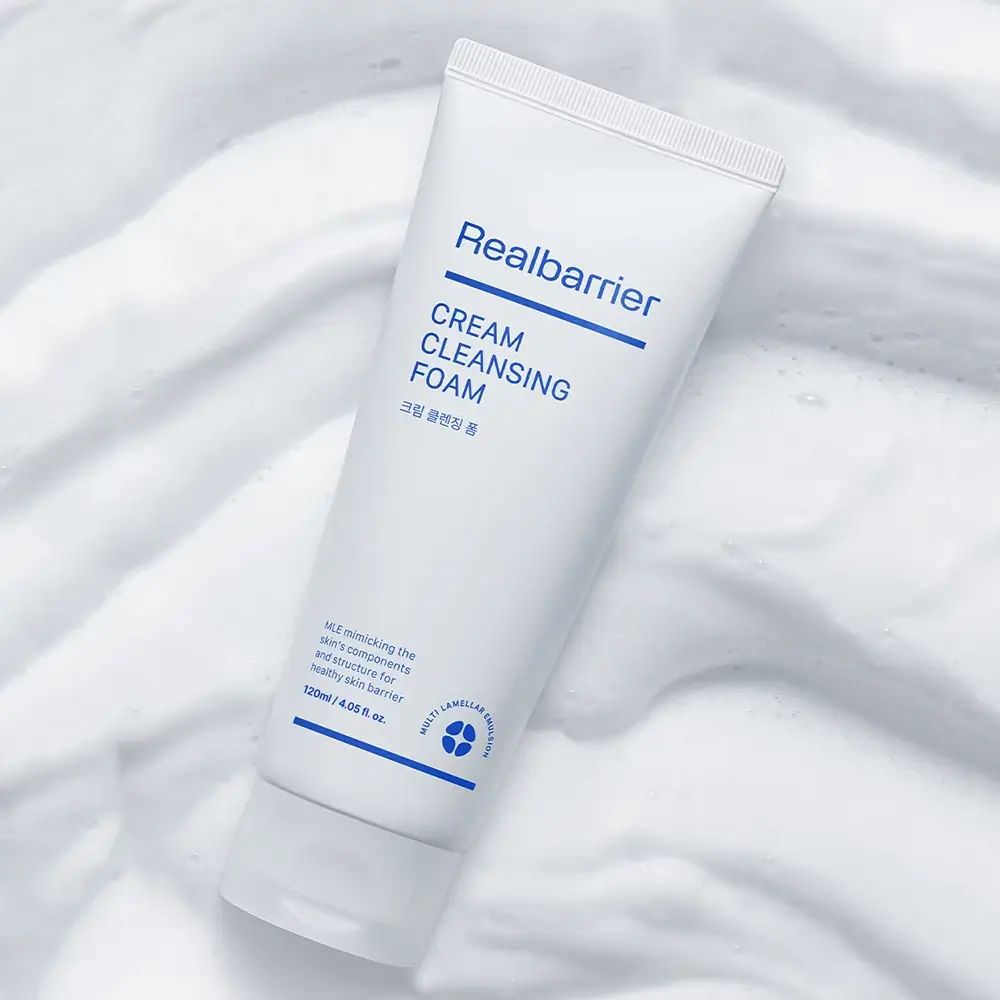 Tubo bianco con testo blu: Realbarrier CREAM CLEANSING FOAM. Su sfondo di schiuma bianca.