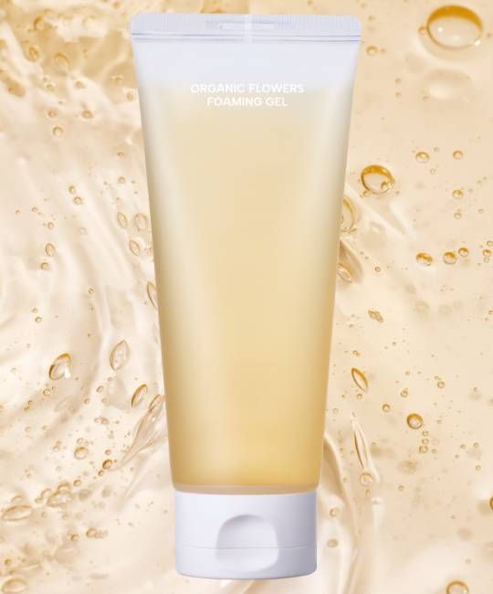 Tubo trasparente con scritta "ORGANIC FLOWERS FOAMING GEL". Liquido giallastro visibile. Tappo bianco. Sfondo con bolle.