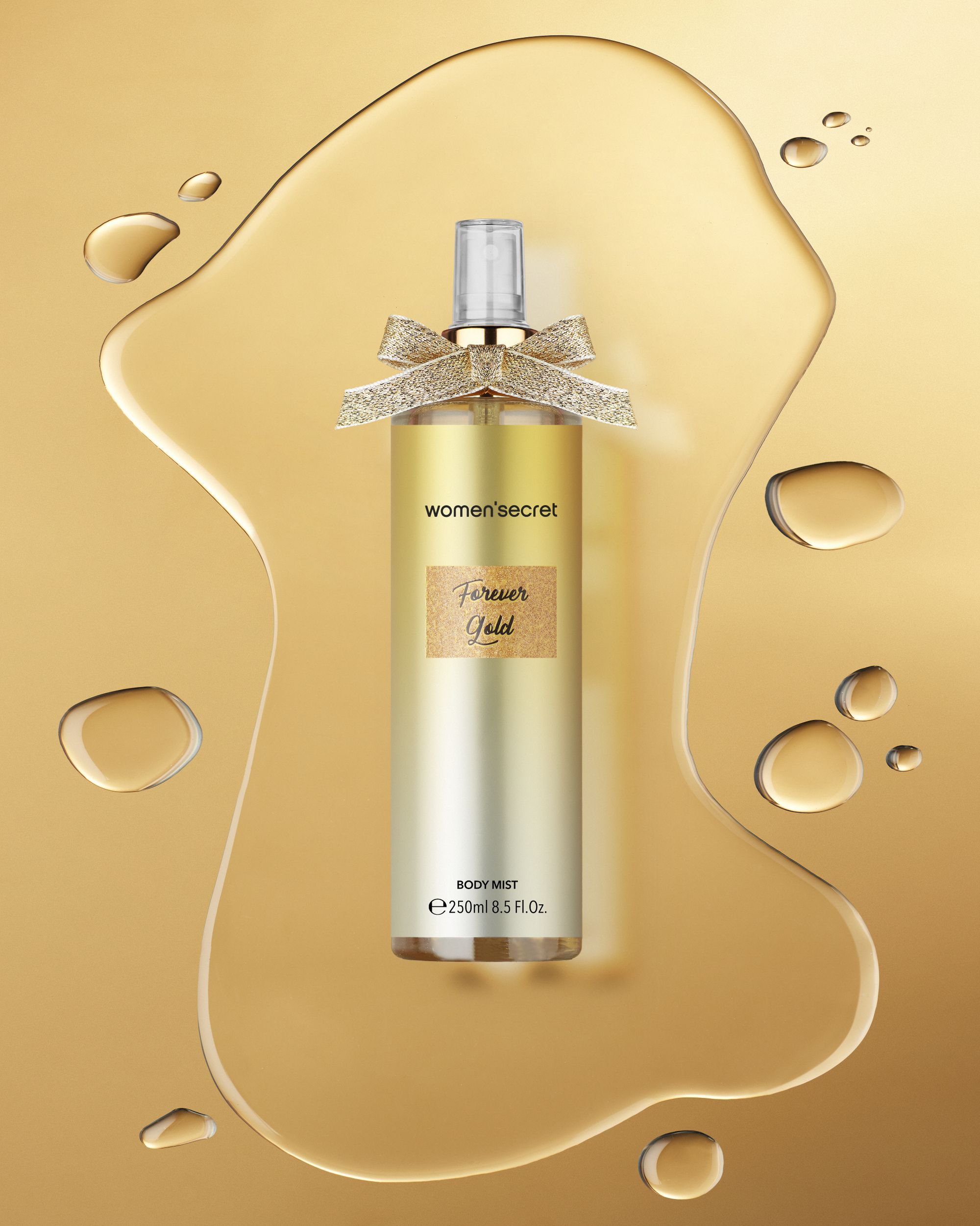 Body mist dorato con fiocco. Scritte: women'secret, Forever Gold, BODY MIST. Gocce di liquido sullo sfondo.