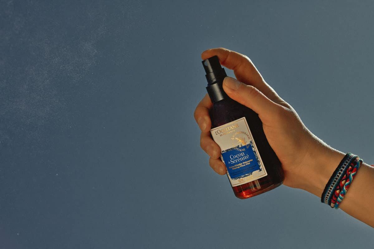 Mano che tiene un flacone marrone con vaporizzatore nero. Nebbia spray visibile. Etichetta con nome del prodotto. L'Occitane Aromachologie Kopfkissen-Nebel Entspannend.