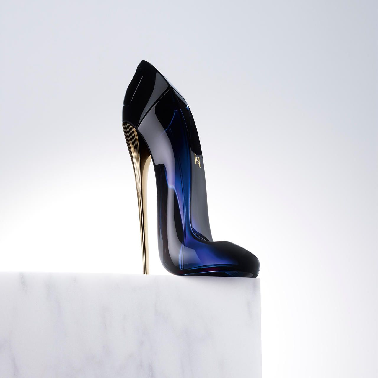 Flacone a scarpa blu con tacco dorato su piedistallo bianco. Ambiente chiaro.