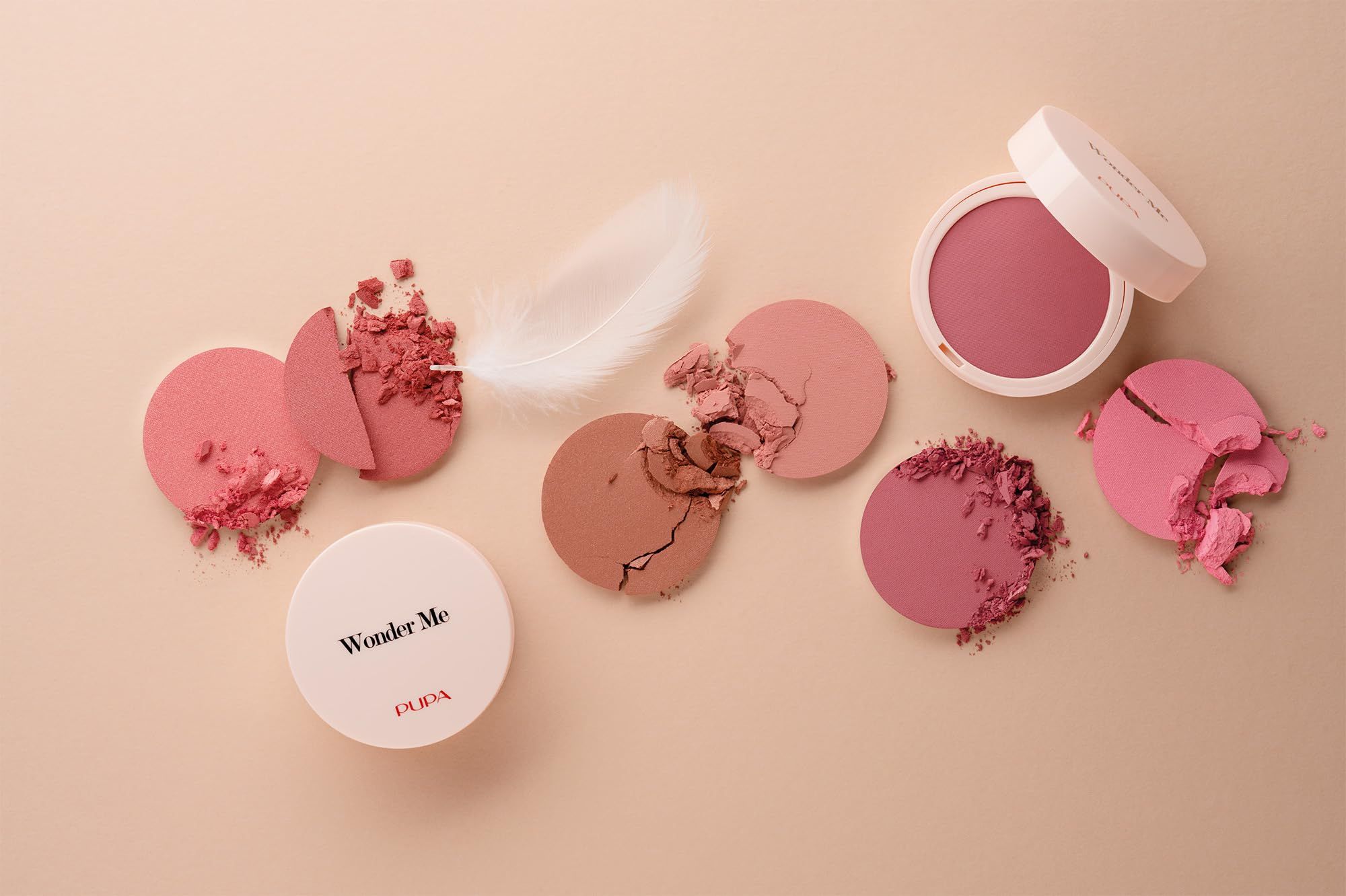 Diverse polveri blush rotte in varie tonalità di rosa. Una polvere è intatta. Piuma accanto.