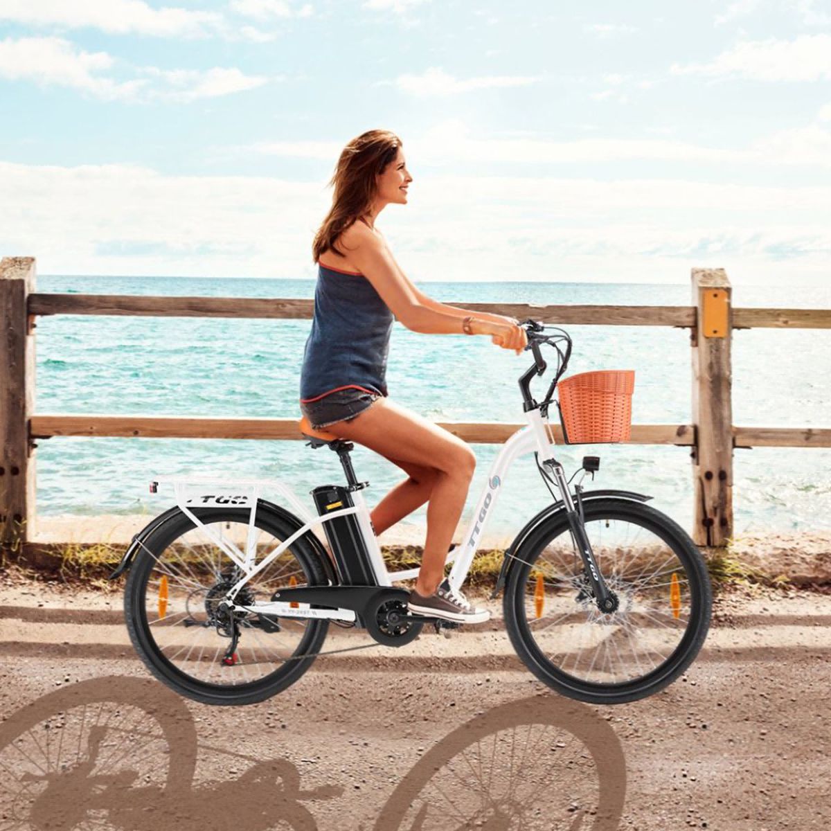 Donna in bicicletta elettrica bianca con cestino. Sella e manopole marroni. Logo TTGO. Sfondo: mare.