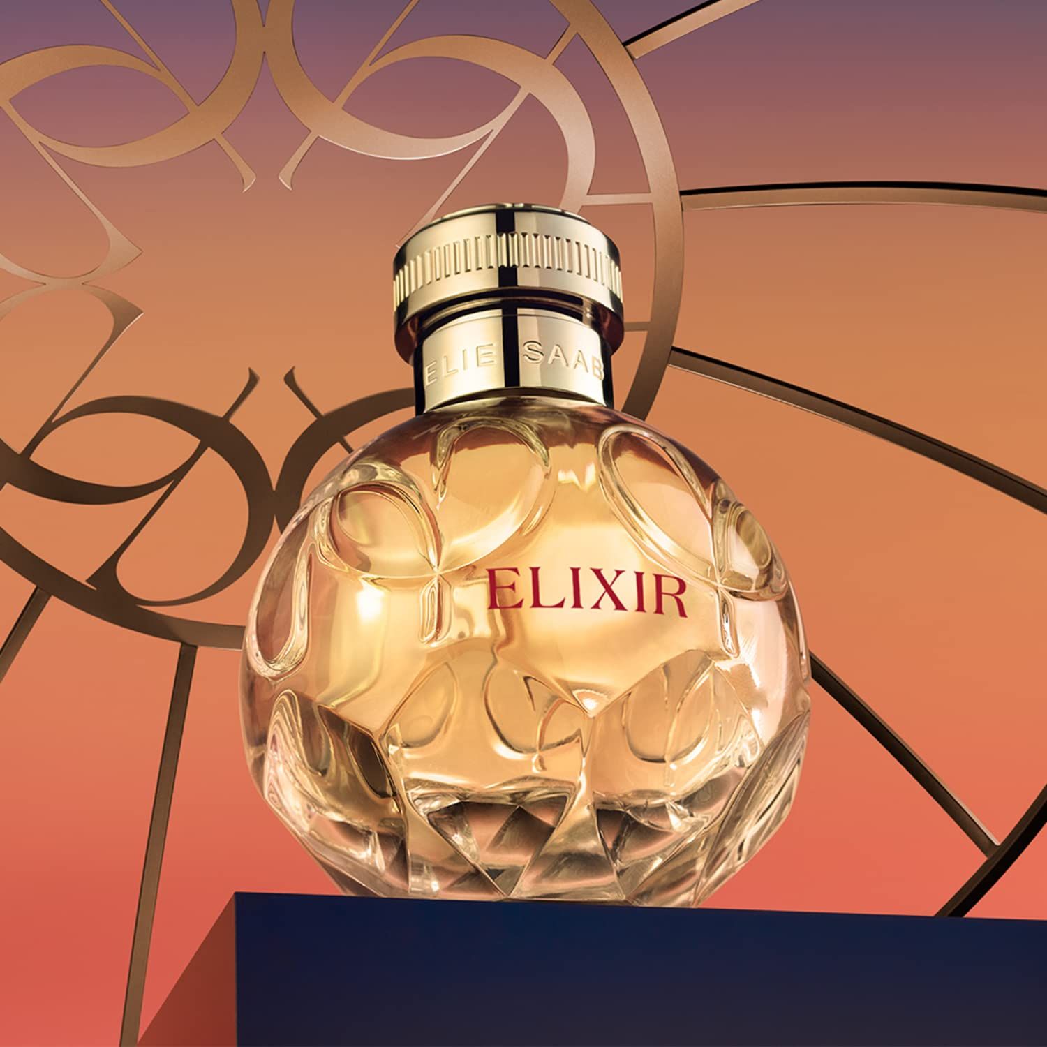 Flacone rotondo con tappo dorato. Scritta "ELIXIR" e "ELIE SAAB". Sfondo con elementi dorati.