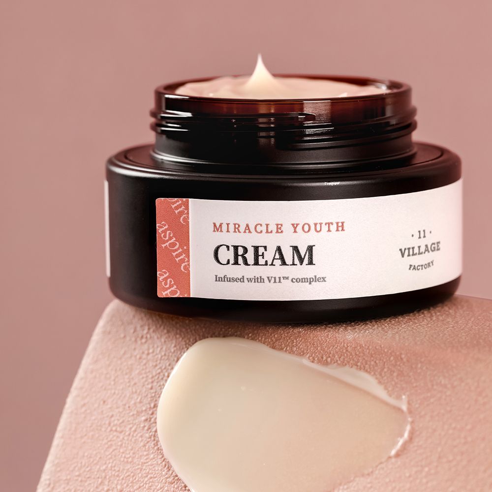 Vasetto aperto con crema. Macchia di crema accanto. Scritta: Miracle Youth Cream, Village 11 Factory. Sfondo rosa.