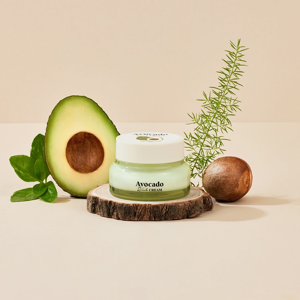 Vasetto di crema su ceppo di legno, accanto a metà avocado, nocciolo e ramo. Scritta "Avocado Rich CREAM".