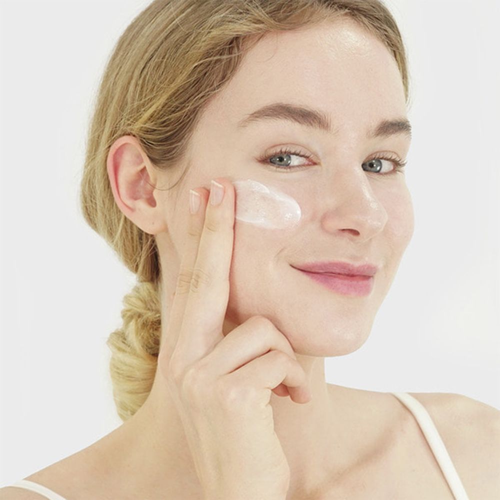 Donna applica crema sul viso. Crema sul dito. Pelle chiara, capelli biondi, top bianco.