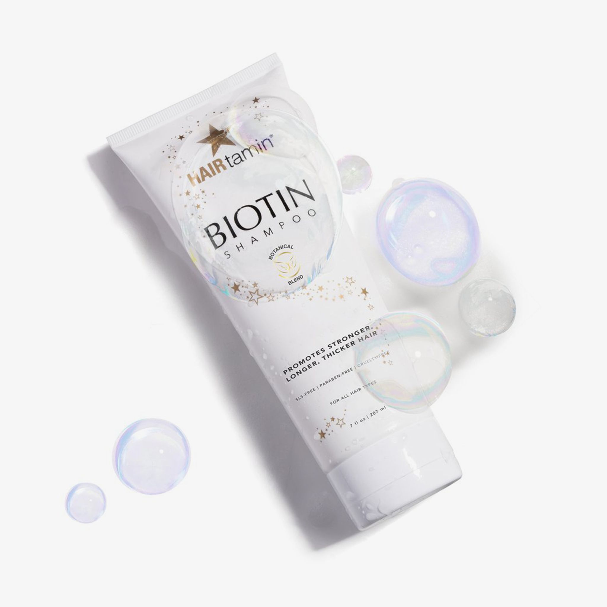 Tubo bianco "HAIRtamin Biotin Shampoo" con bolle di sapone. Stelle e scritta "Botanical Blend". Testo: "Supports stronger, longer, thicker hair".