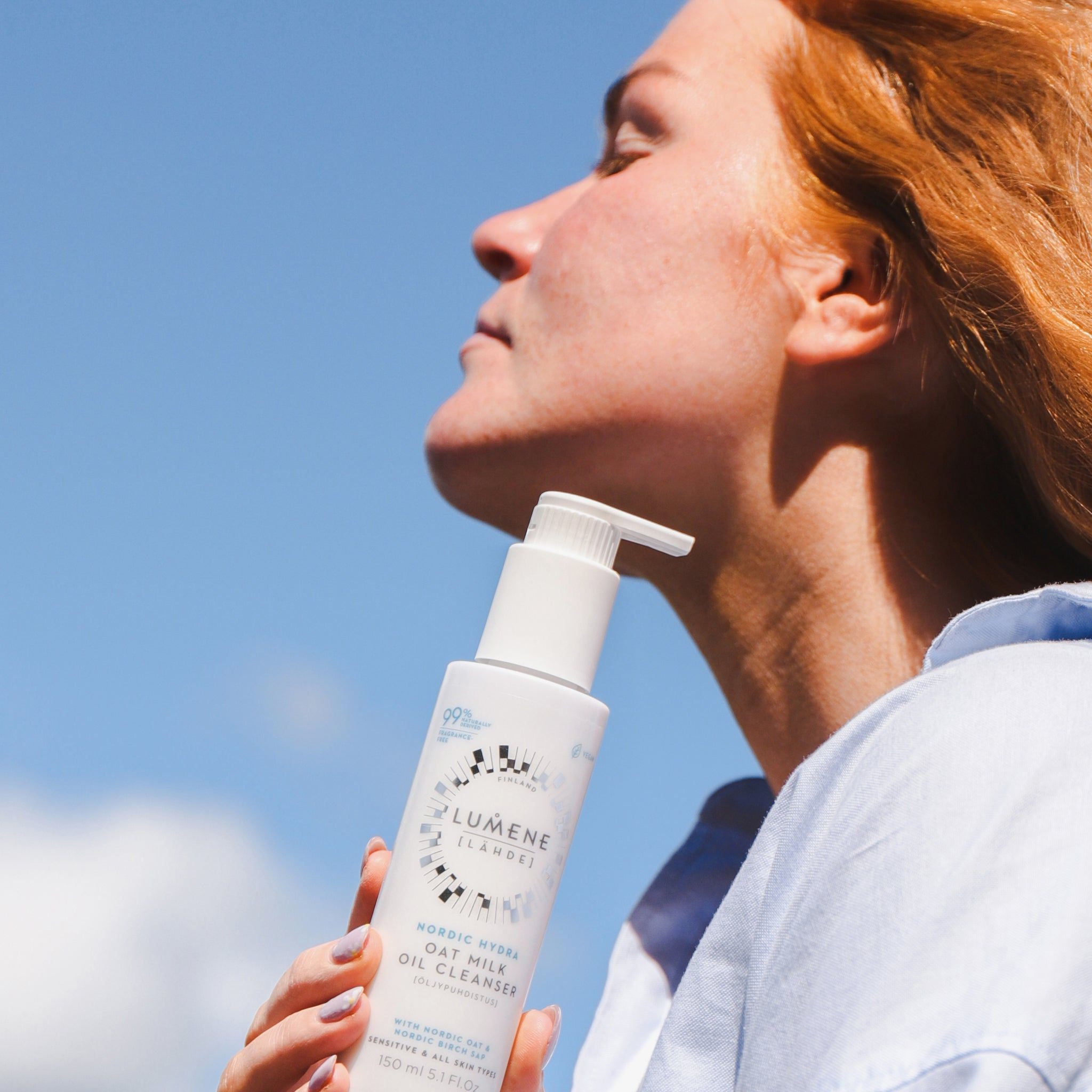 Donna tiene un flacone bianco con dosatore. Scritta: Lumene Nordic Hydra Oat Milk Oil Cleanser. 99% naturale. Cielo blu.
