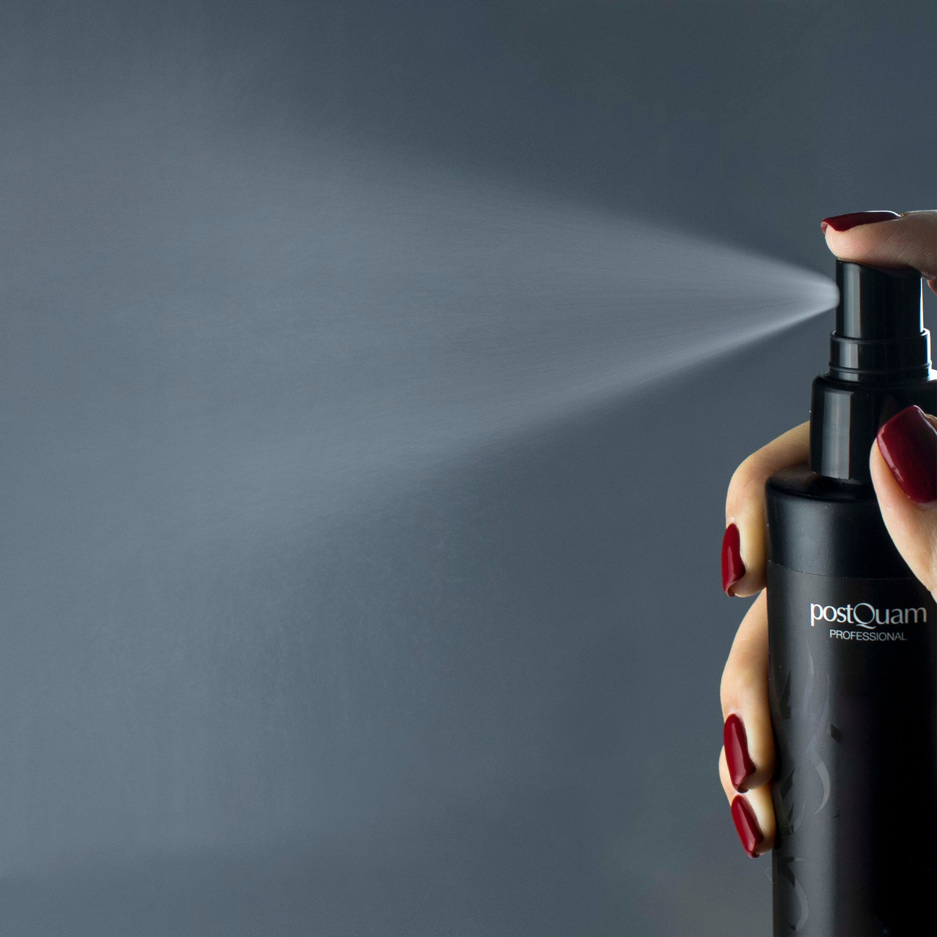 Mano che spruzza da flacone nero con "postQuam PROFESSIONAL". Unghie rosse. Nebbia di spruzzo visibile.