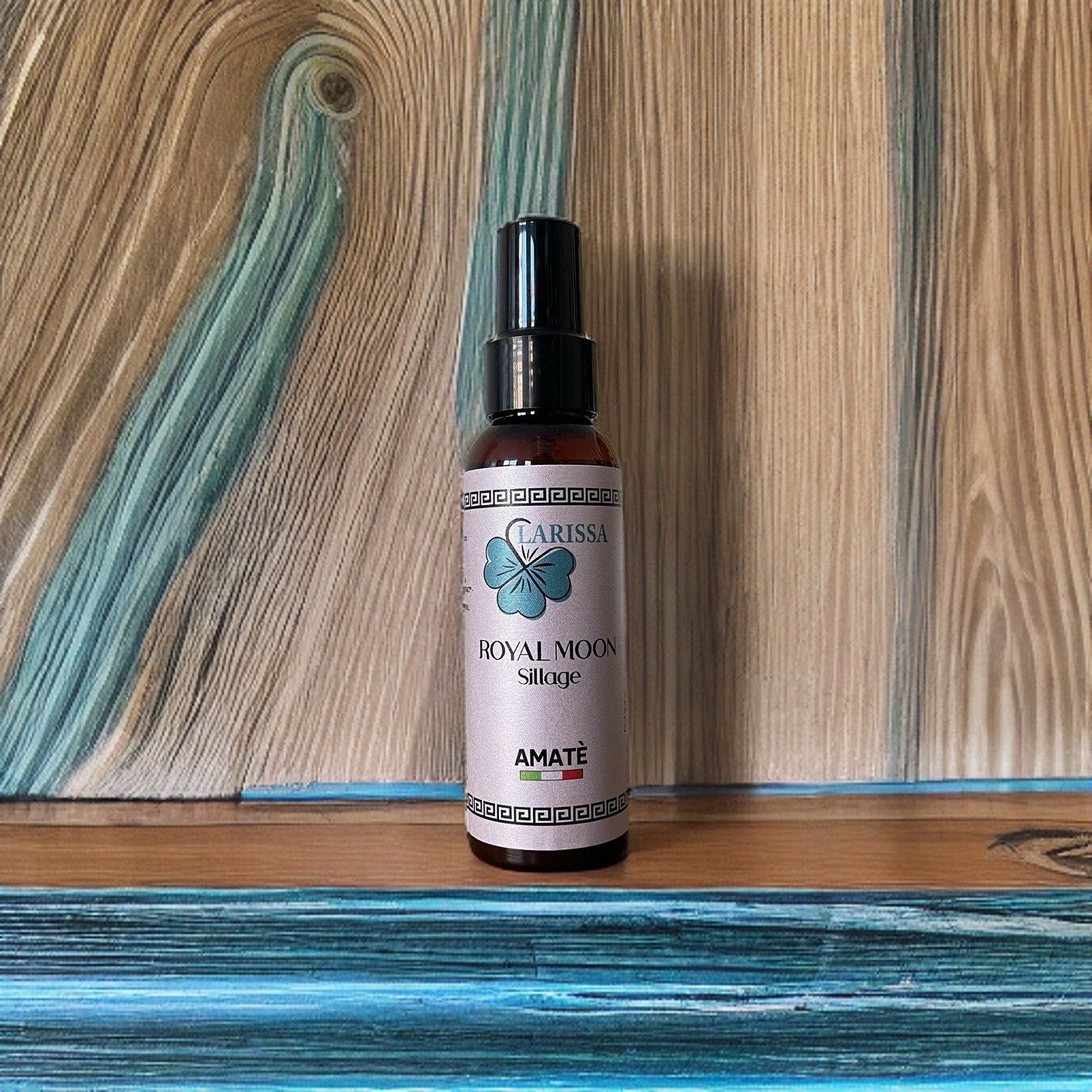 Flacone marrone con vaporizzatore nero. L'etichetta riporta "Larissa Royal Moon Sillage" e "Amatè". Appoggiato su una tavola di legno.