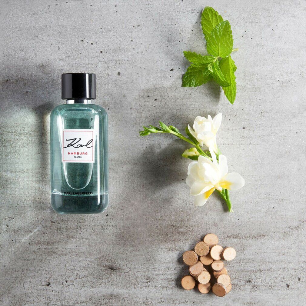 Karl Lagerfeld Hamburg Alster E.d.T. Nat. Spray. Flacone con tappo nero, accanto a fiori e pezzi di legno.
