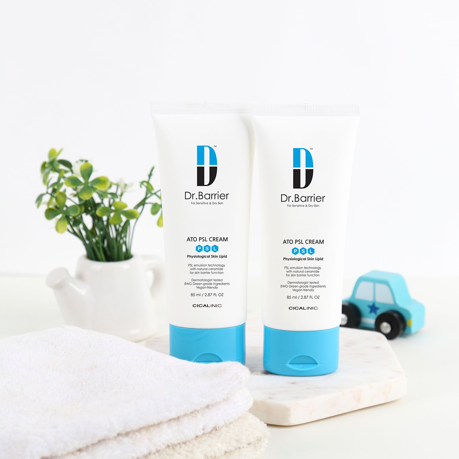 Due tubi di Dr.Barrier ATO PSL Cream. Su una superficie con pianta, asciugamano e macchinina. Schema colori blu e bianco.