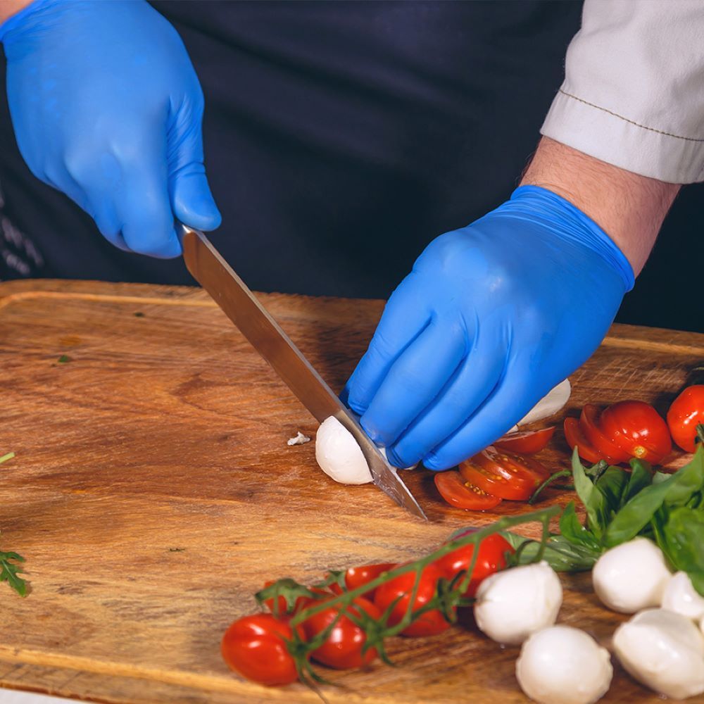 Mani con guanti blu tagliano cibo su tagliere in legno. Pomodori e mozzarella.