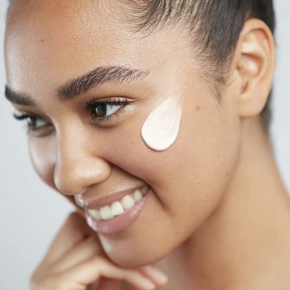 Viso di donna sorridente. Una goccia di crema sulla guancia. Pelle chiara, sopracciglia scure.