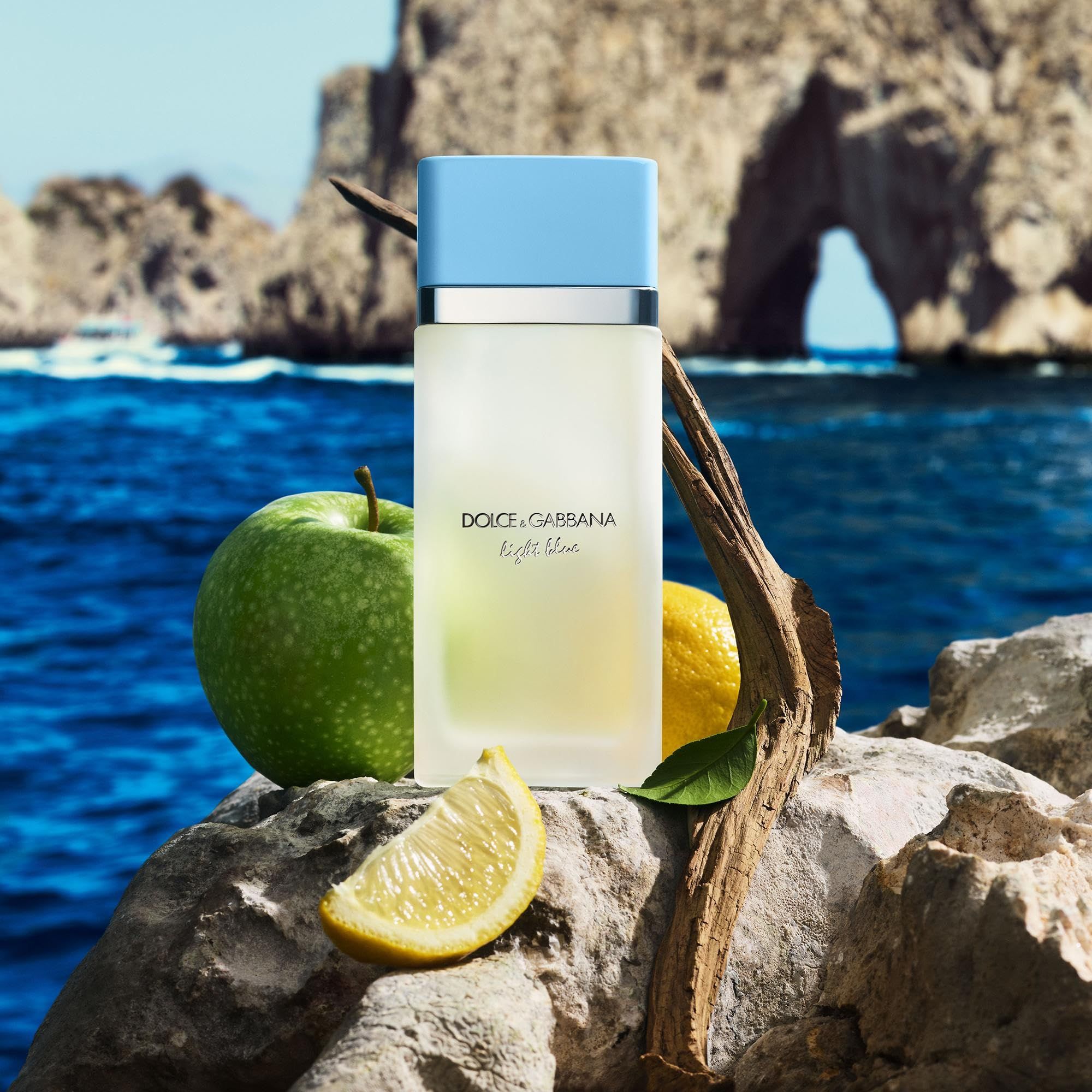 Flacone su pietra. Mela, limone e ramo accanto. Sfondo: mare e rocce. Scritta: Dolce & Gabbana Light Blue.