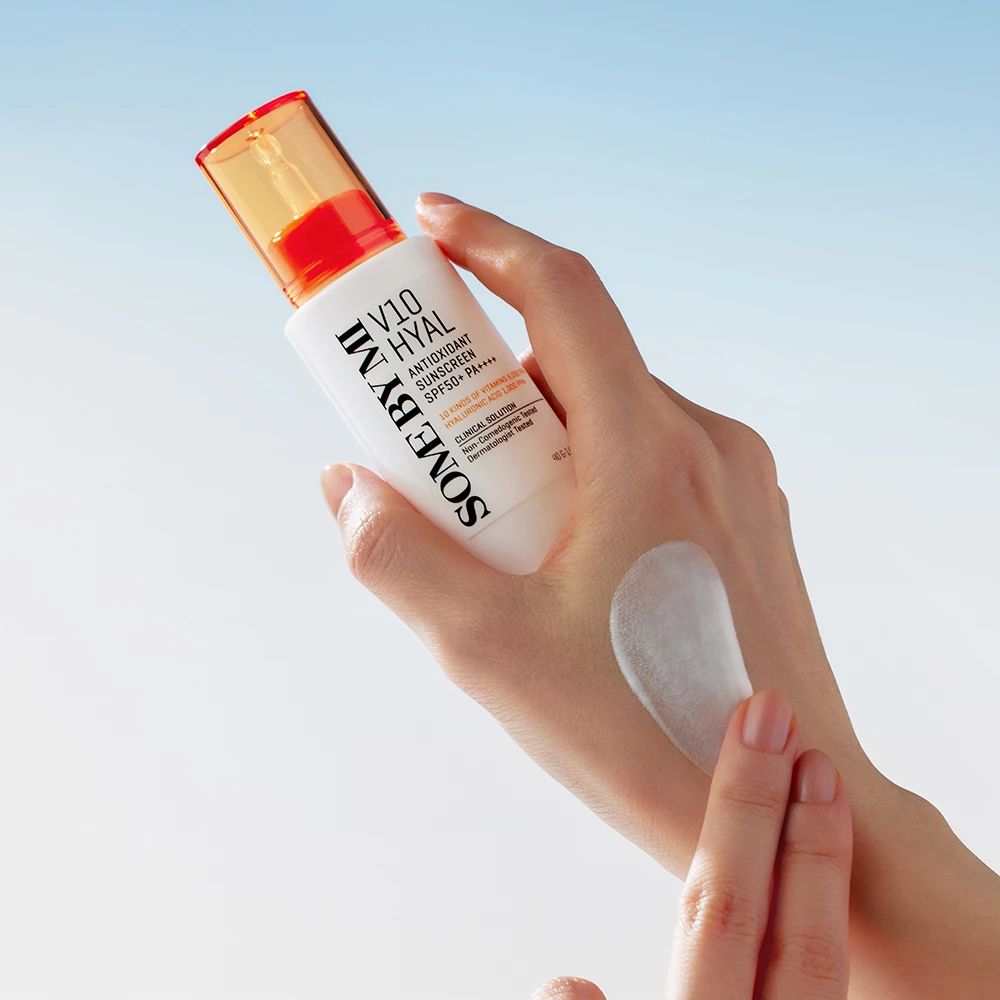 Flacone bianco con tappo arancione. Prodotto applicato sulla mano. Testo: SOME BY MI, V10 HYAL Antioxidant Sunscreen SPF50+.
