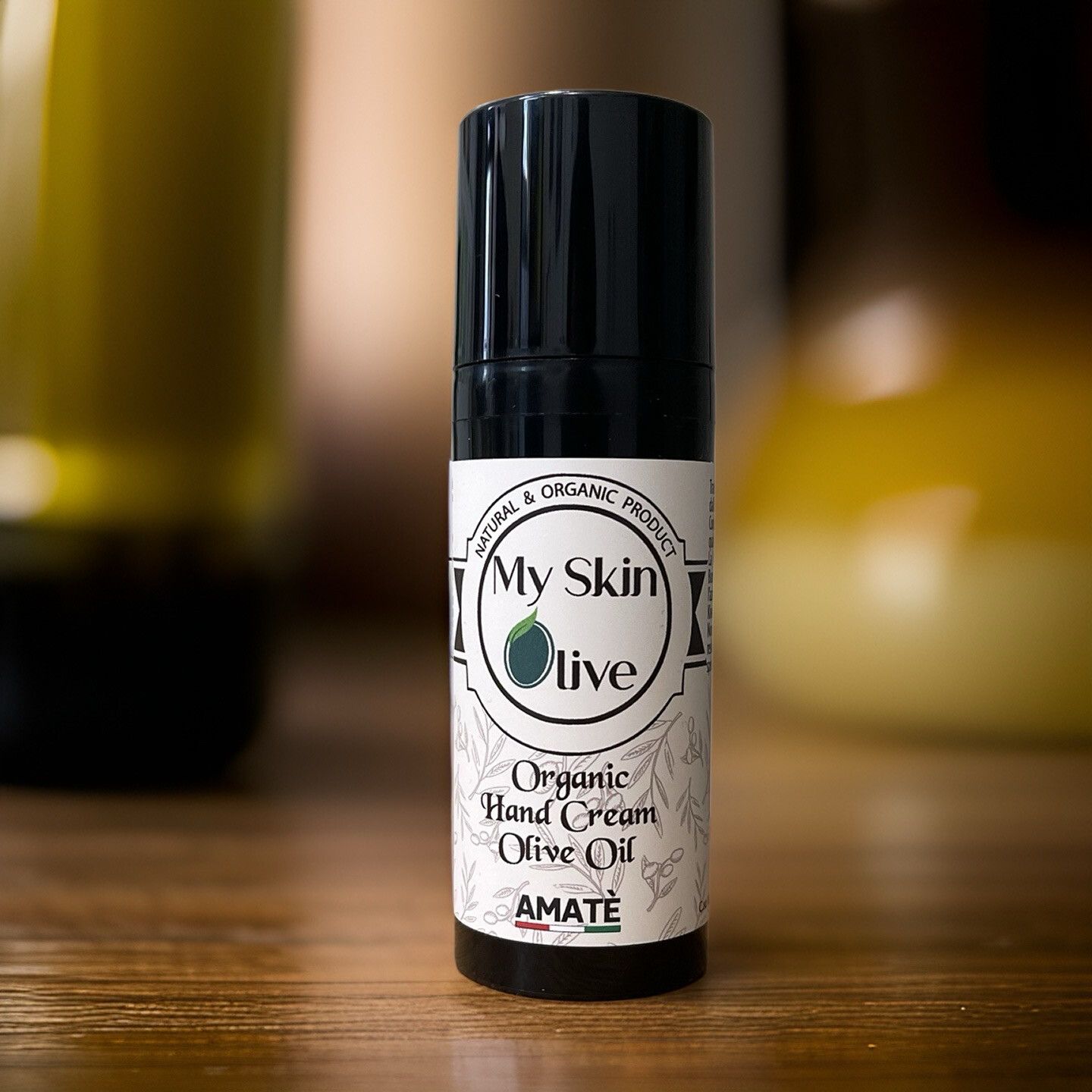 Flacone nero su sfondo sfocato. Scritta: My Skin Olive, Organic Hand Cream Olive Oil, Amatè.