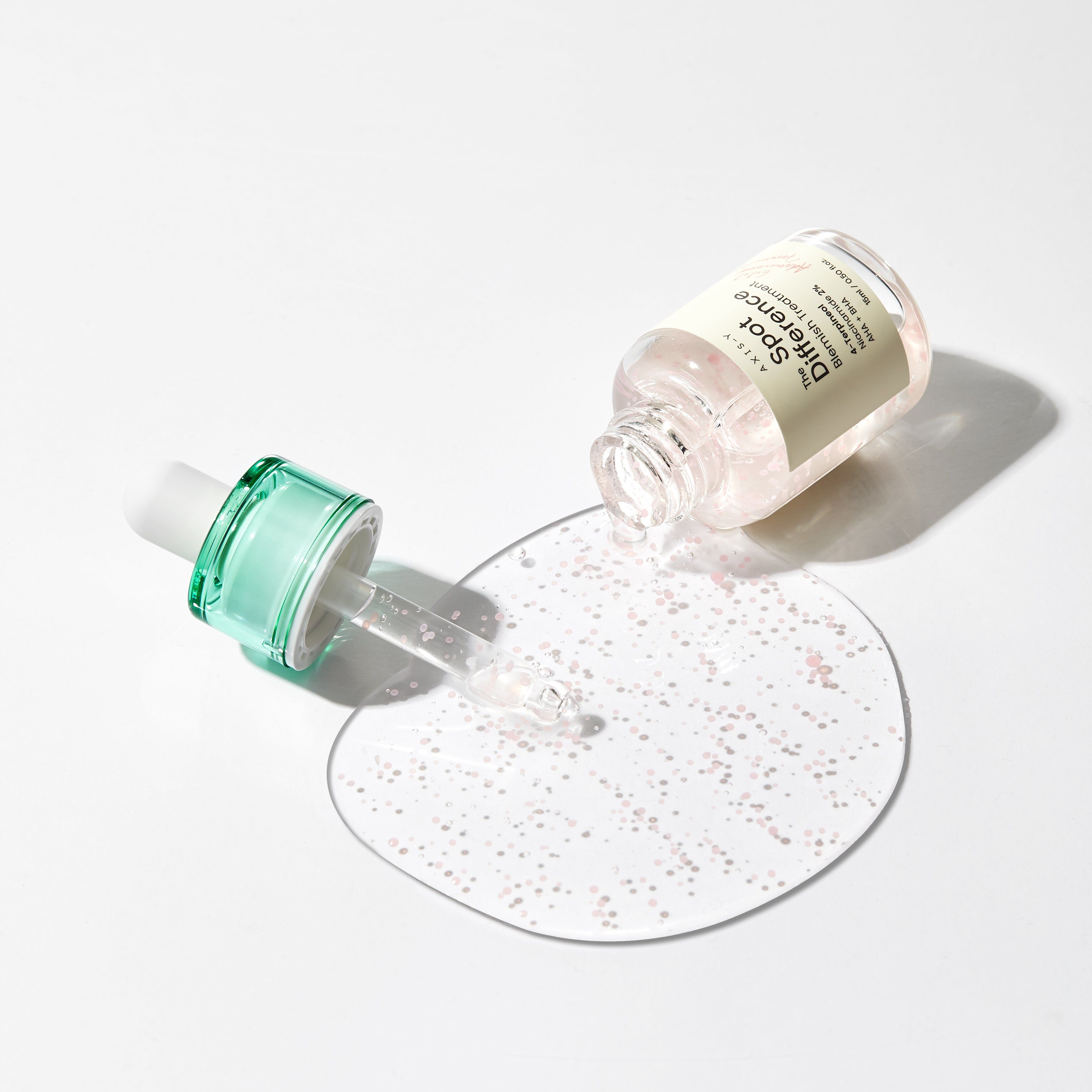 Flacone del prodotto sdraiato, liquido che fuoriesce. Pipetta accanto. Piccole sfere rosa sulla superficie. Sfondo bianco.