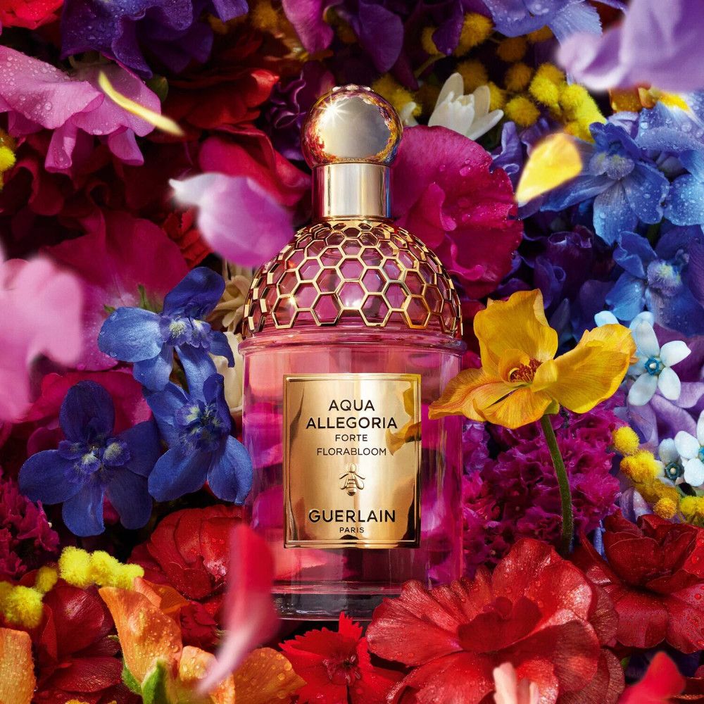 Flacone di vetro rosa con tappo dorato, circondato da fiori colorati. Scritta: Aqua Allegoria Forte Florabloom, Guerlain Paris.
