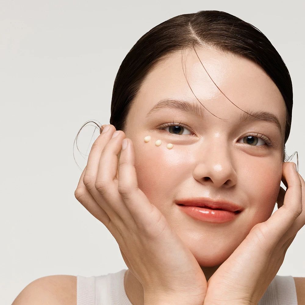 Ritratto di una donna con punti di crema sotto gli occhi. Sorride. Mani sul viso.