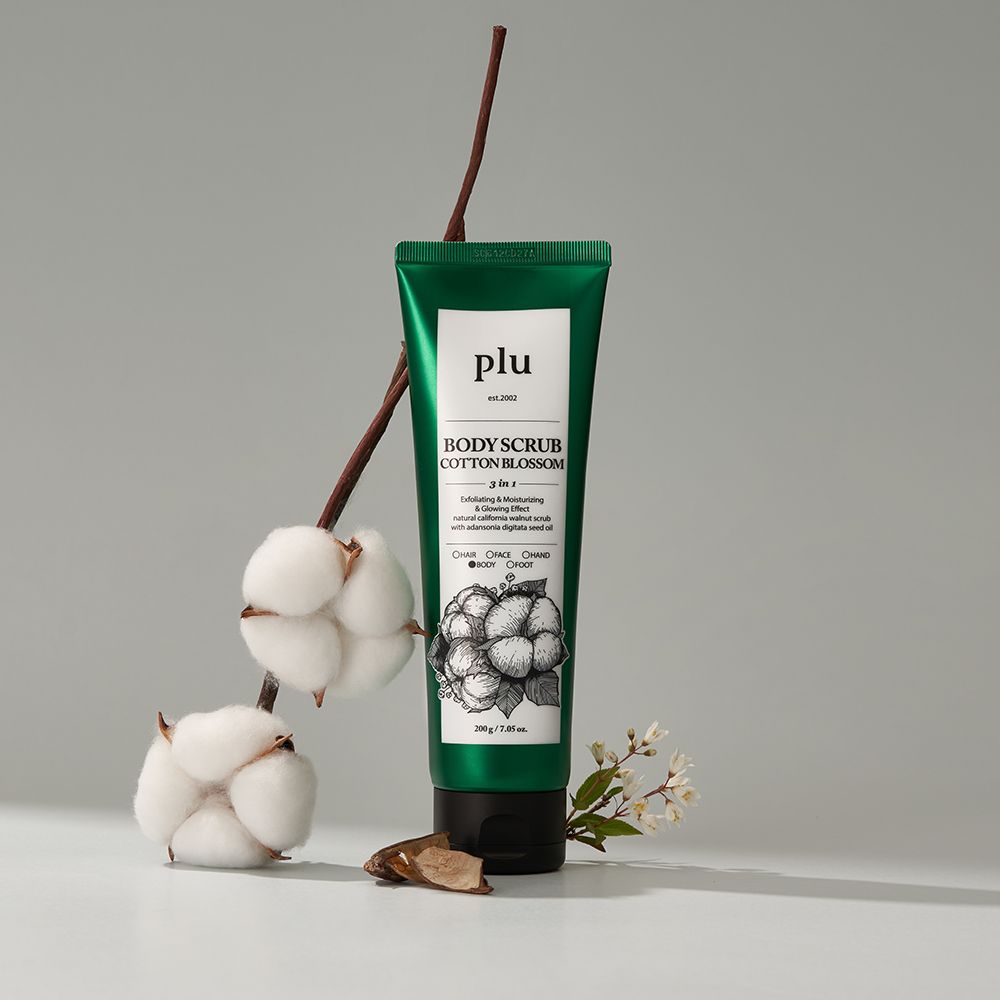 Tubo verde con etichetta bianca. Scritta: PLU Body Scrub Cotton Blossom. Ramo di cotone e fiori accanto al tubo, su sfondo chiaro.