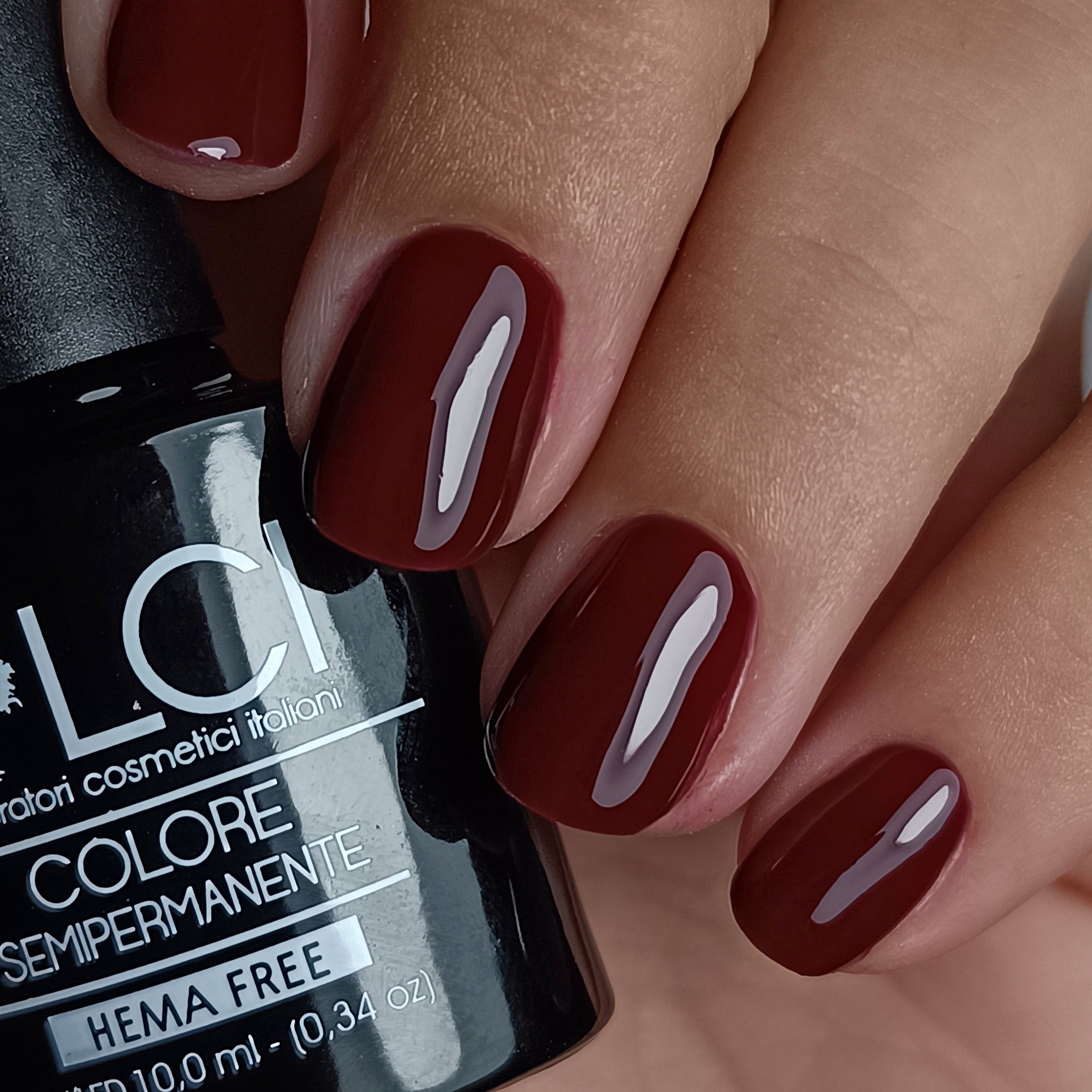 Mano con unghie smaltate. Smalto rosso scuro. Flacone con scritta: LCI, Colore Semipermanente, HEMA FREE.