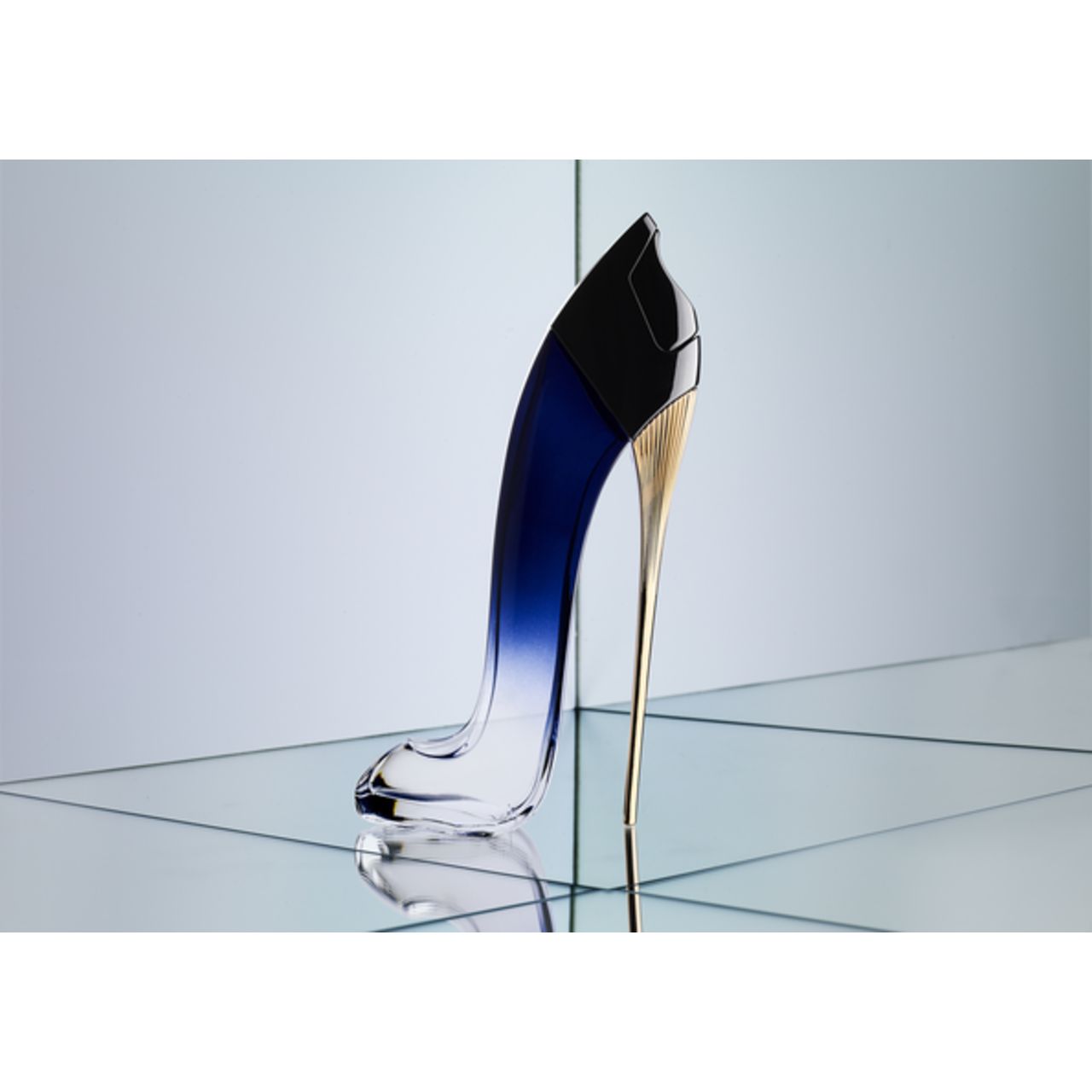 Flacone di profumo a forma di tacco alto. Sfumatura nero-blu, tacco dorato. Posizionato su superficie vetrata.