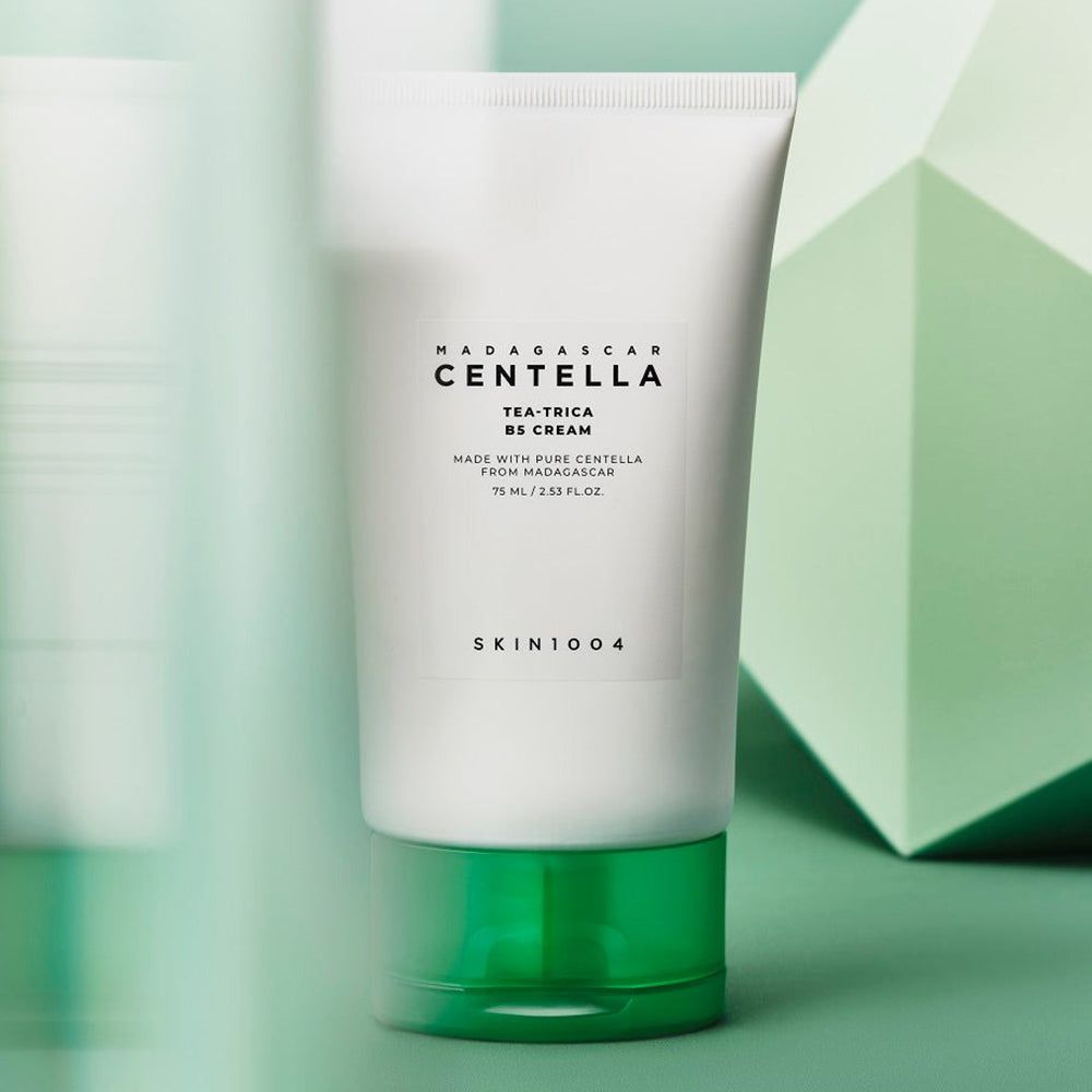 Tubo bianco con base verde. Scritta: MADAGASCAR CENTELLA, TEA-TRICA B5 CREAM, SKIN1004. Sfondo: forme geometriche.