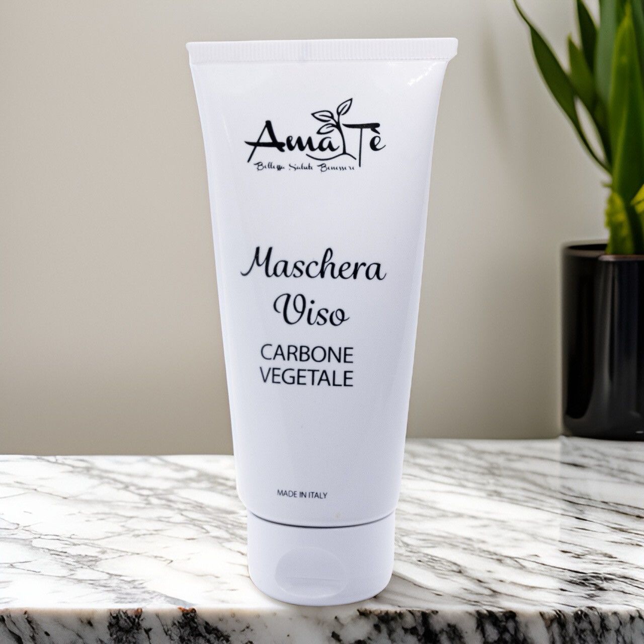 Tubo bianco Amatè Maschera Viso Carbone Vegetale su superficie marmorea. Pianta sullo sfondo.