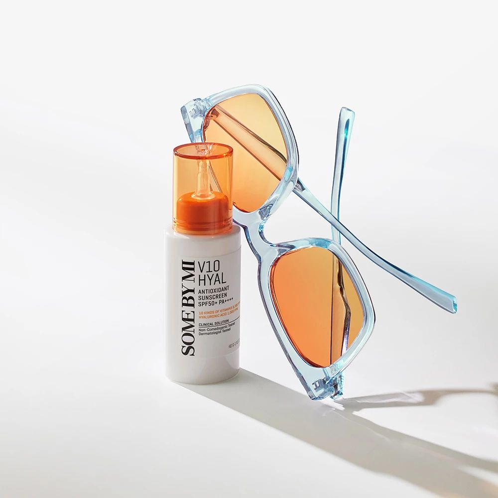 Flacone bianco con tappo arancione accanto a occhiali da sole blu. Testo: V10 HYAL Antioxidant Sunscreen SPF50+.