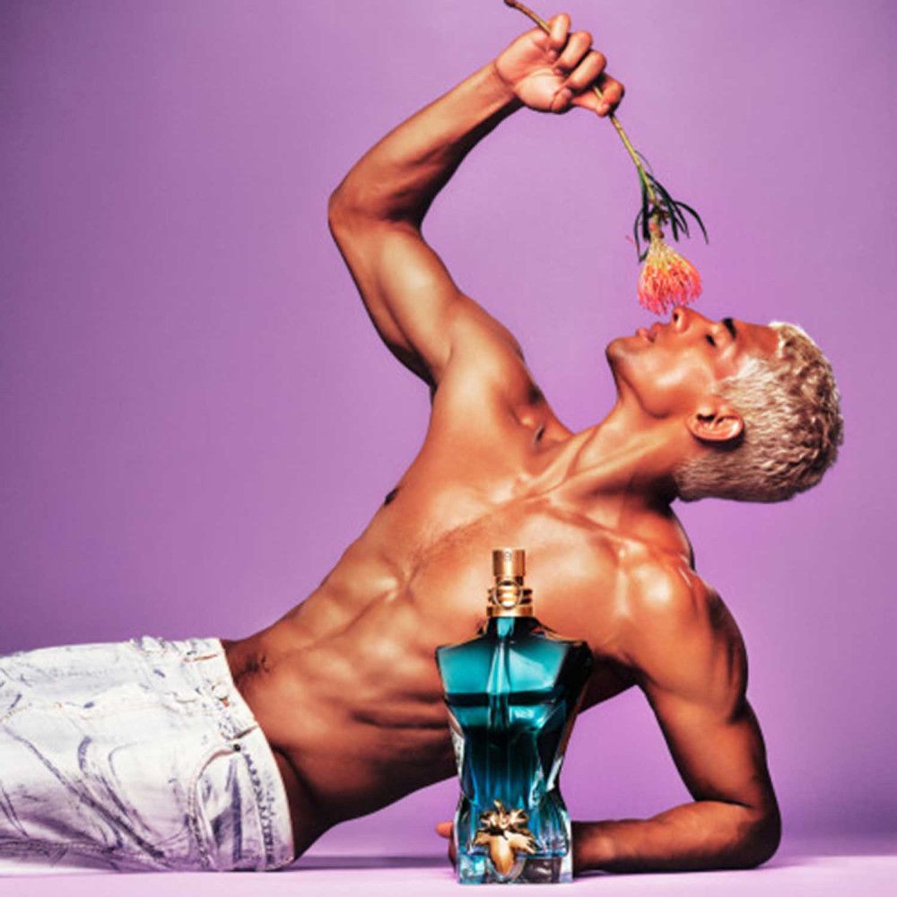 Uomo biondo con fiore. Flacone Jean Paul Gaultier "Le Beau" a forma di torso. Su sfondo viola.