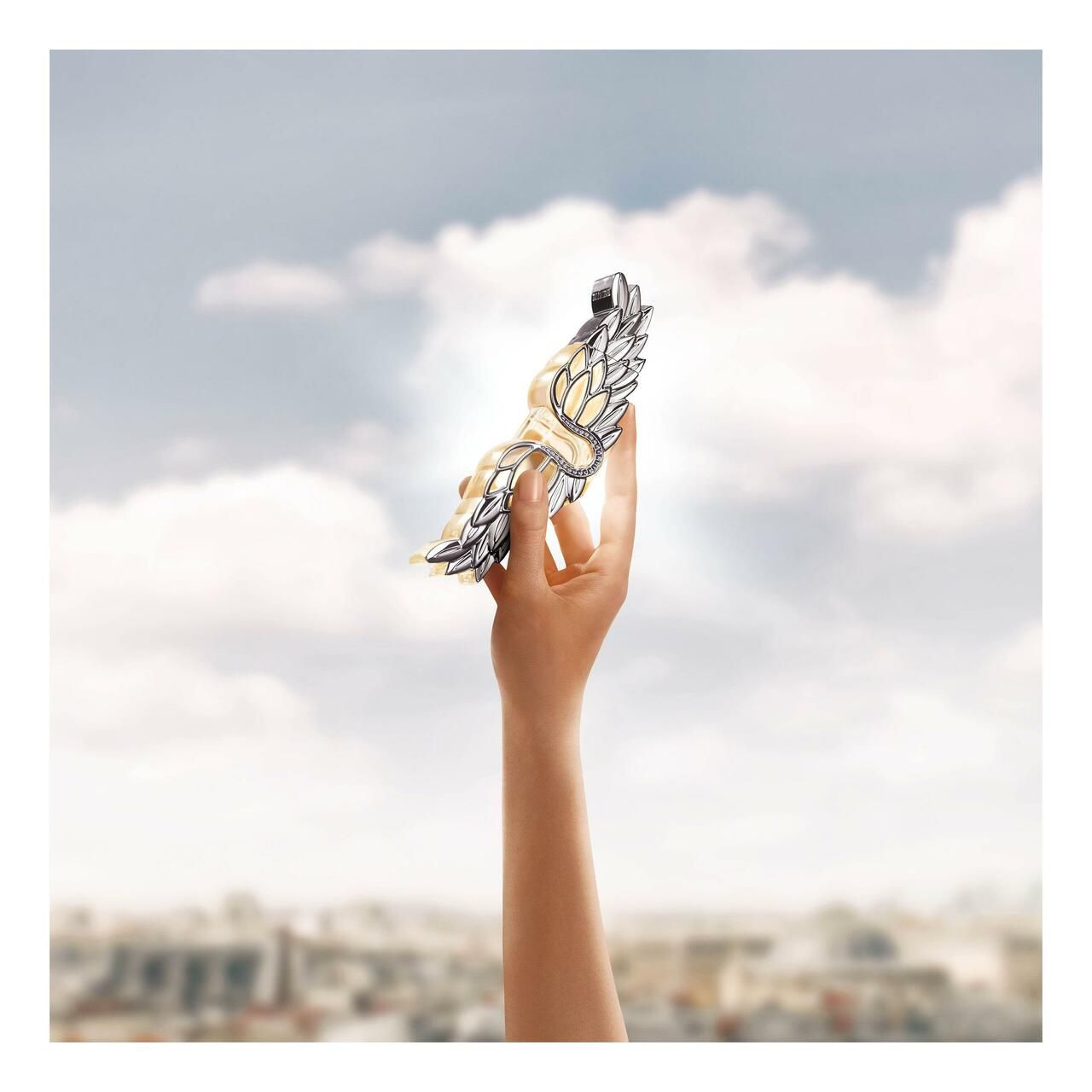 Mano che tiene una decorazione ad ali argentate davanti a un panorama urbano e a un cielo sfocati.